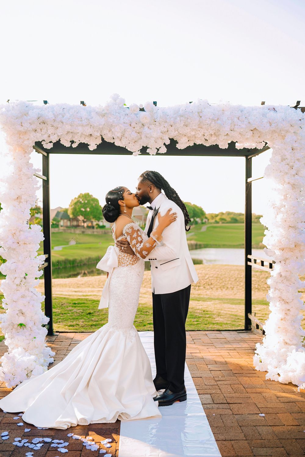 Bride and groom share a first kiss under a white floral arch at an outdoor wedding ceremony.