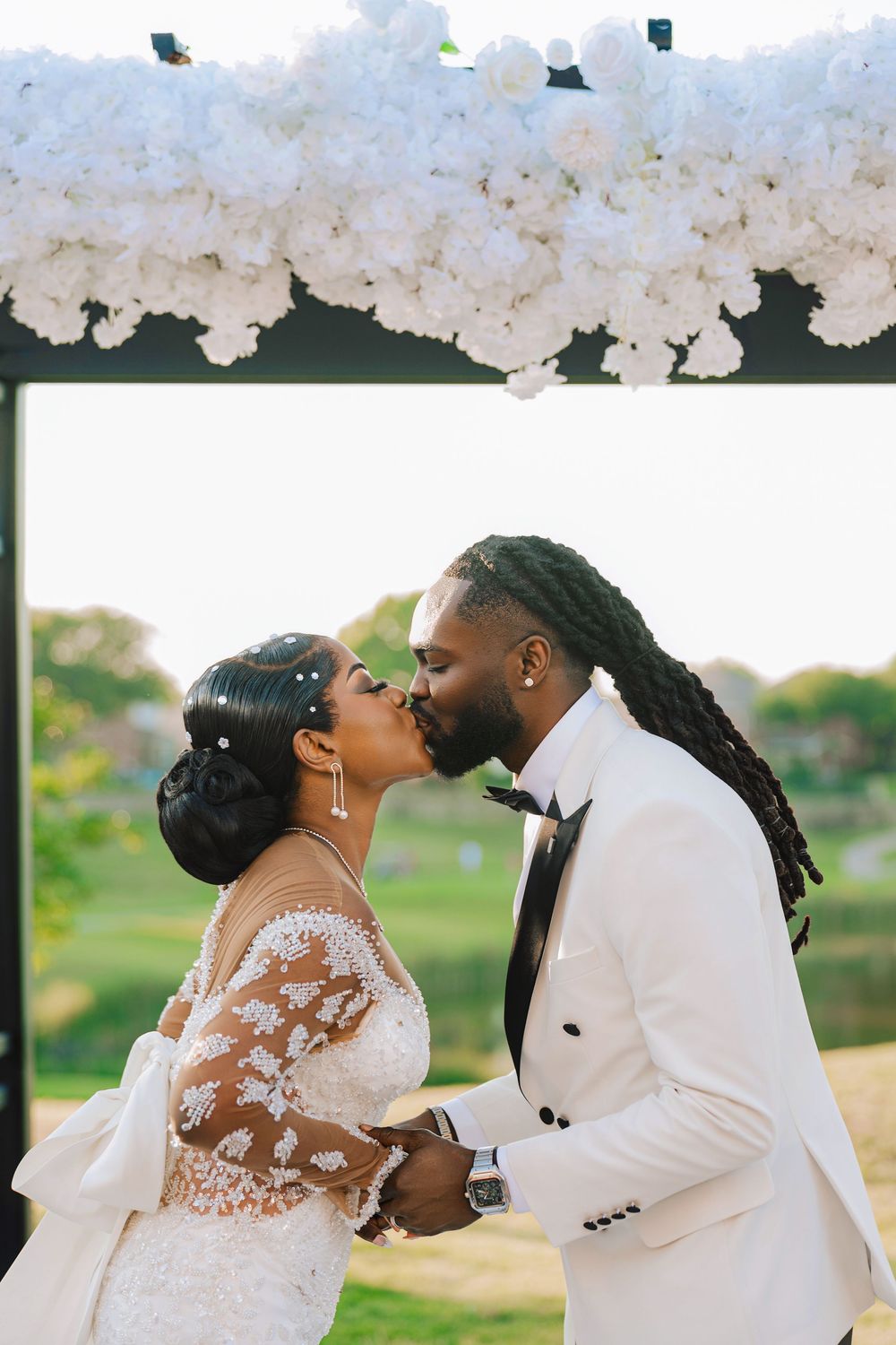 Bride and groom sharing a first kiss under a white floral arch, groom in white tuxedo and bride in lace gown.