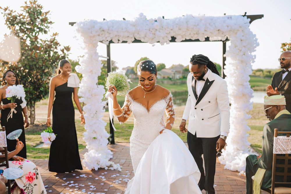 Newlyweds walk back down aisle under white floral arch after outdoor wedding ceremony.