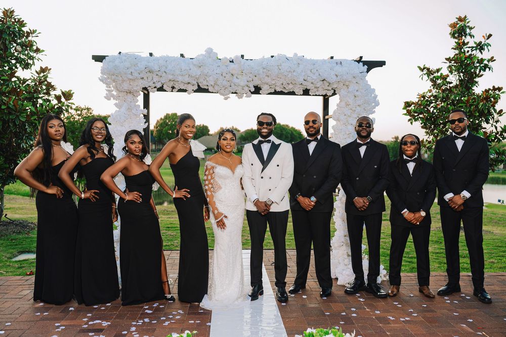 Wedding party posing outdoors, bridesmaids in black gowns and groomsmen in black suits with groom in white tuxedo.