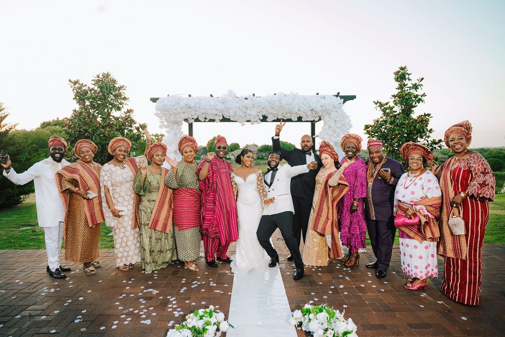 Joyful wedding party celebrating in colorful Nigerian traditional attire in front of a white floral arch outdoors.