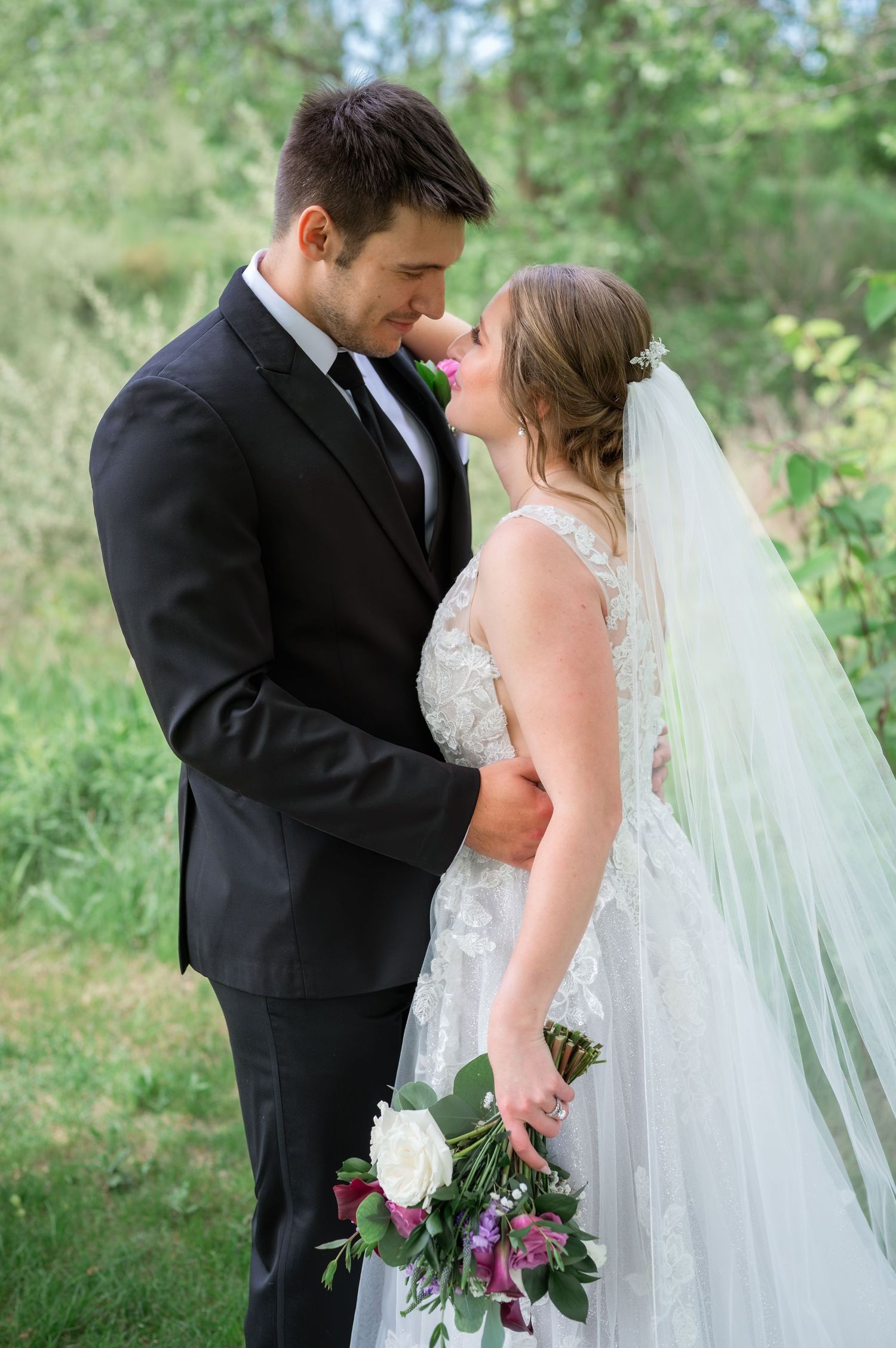 Bride and groom share a romantic nose-to-nose moment outdoors, she holds a white and purple bouquet wearing a lace gown with veil.