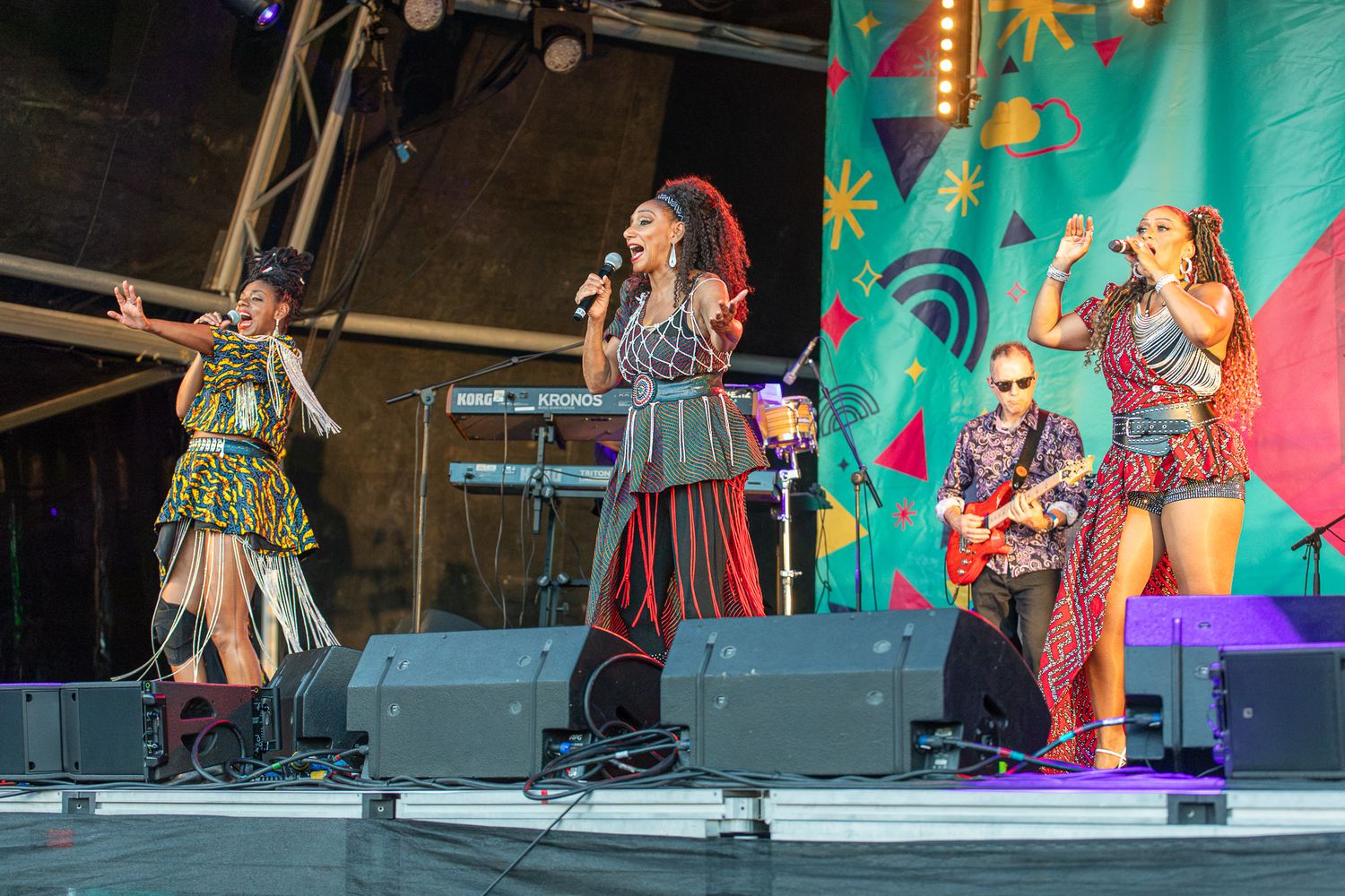 Four female performers singing and playing guitar on a colorful outdoor festival stage.