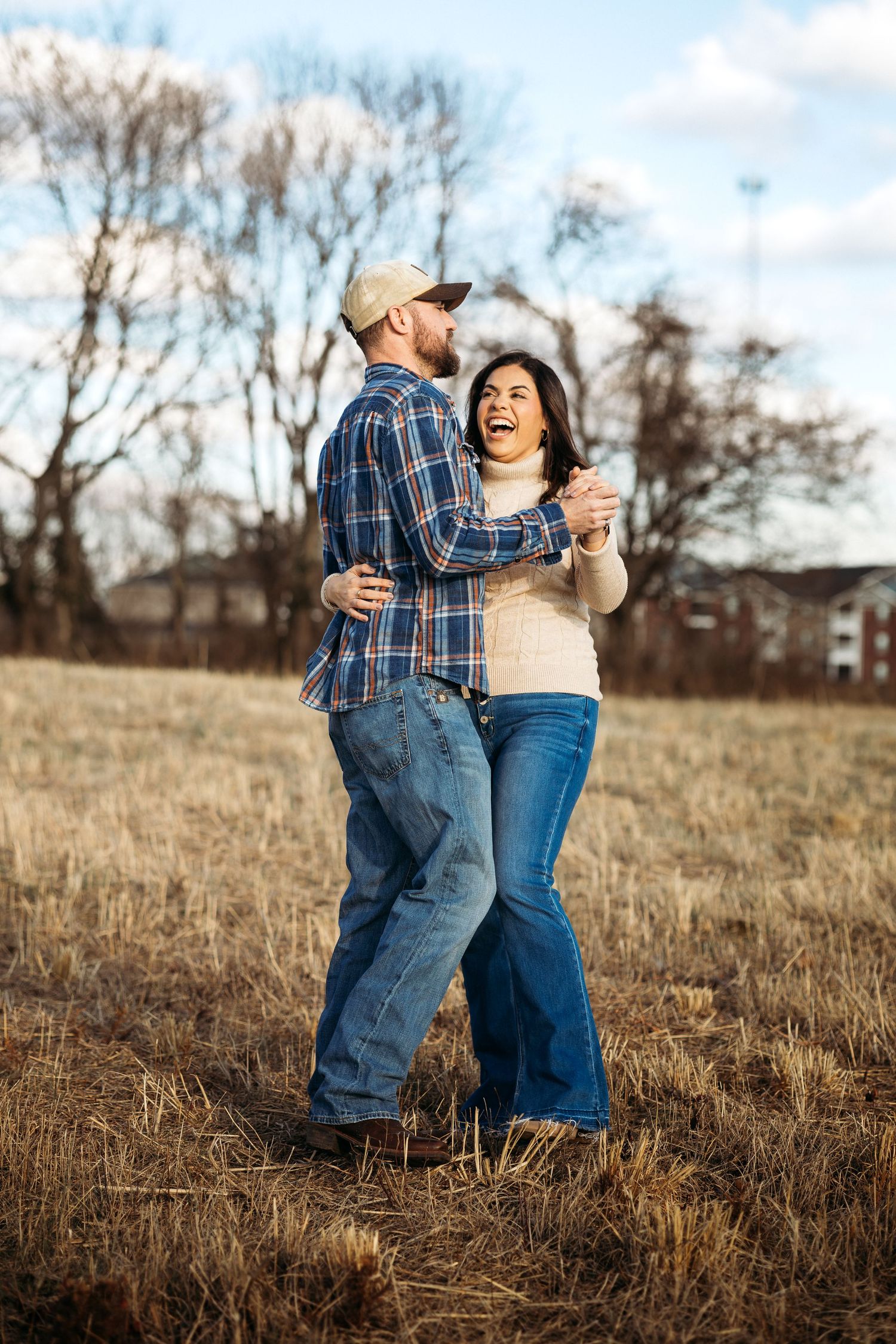 A happy couple dances together in an open field, wearing jeans and flannel, with bare trees in the background.