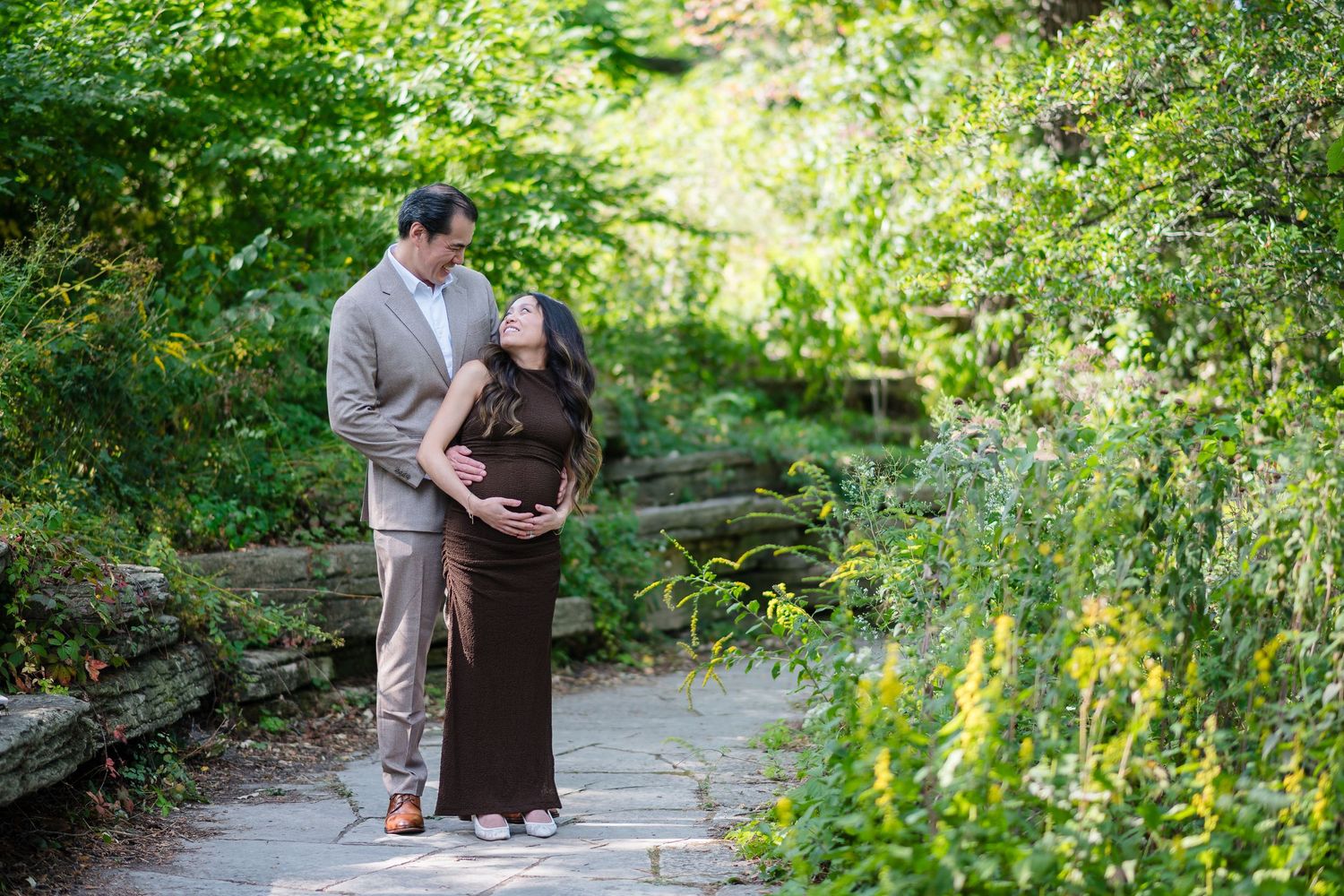 Couple poses on garden path, man in gray suit embraces pregnant woman in brown dress surrounded by lush greenery.