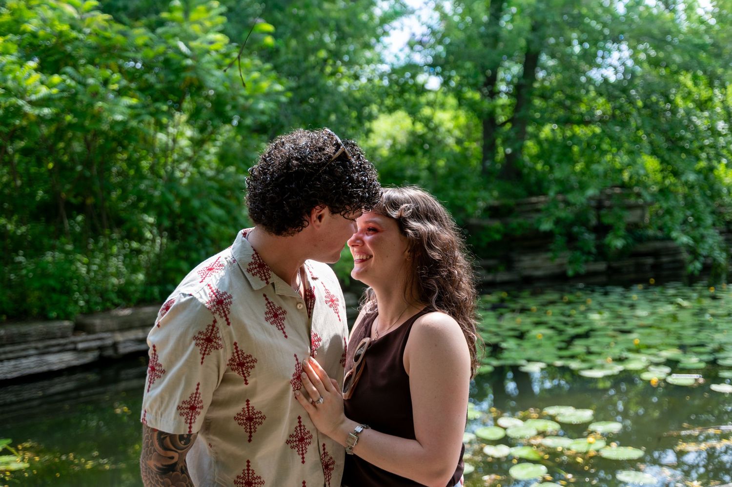 Couple sharing a romantic moment near a lily pad pond surrounded by lush green trees on a sunny day.