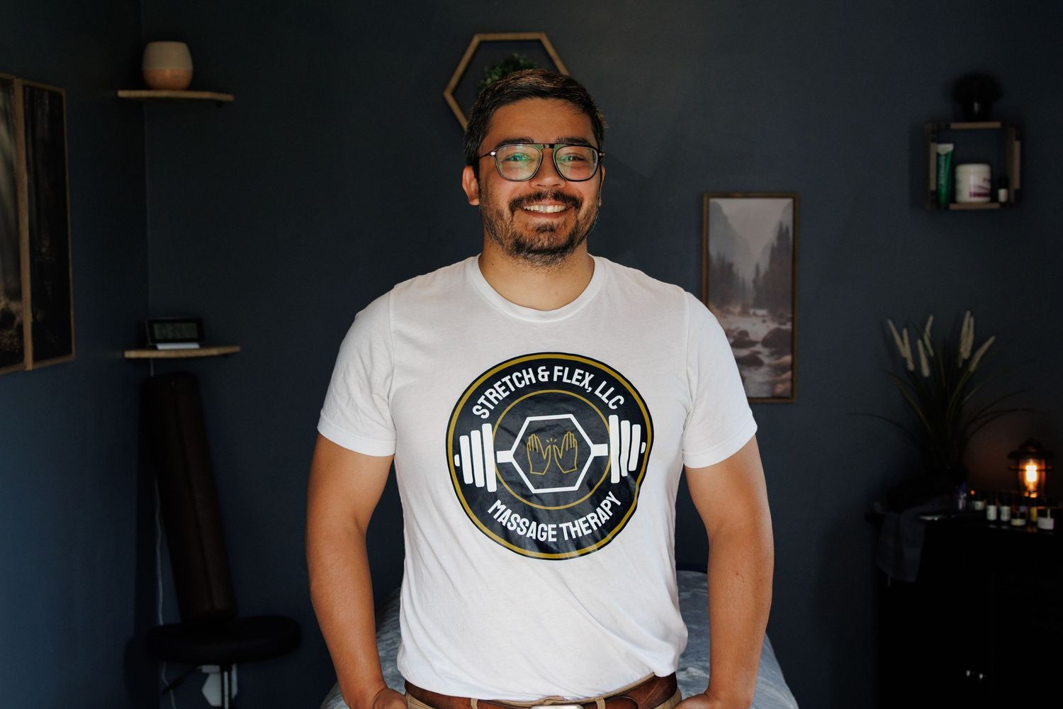 Smiling man wearing a white Stretch & Flex LLC massage therapy t-shirt, standing against a dark teal wall.