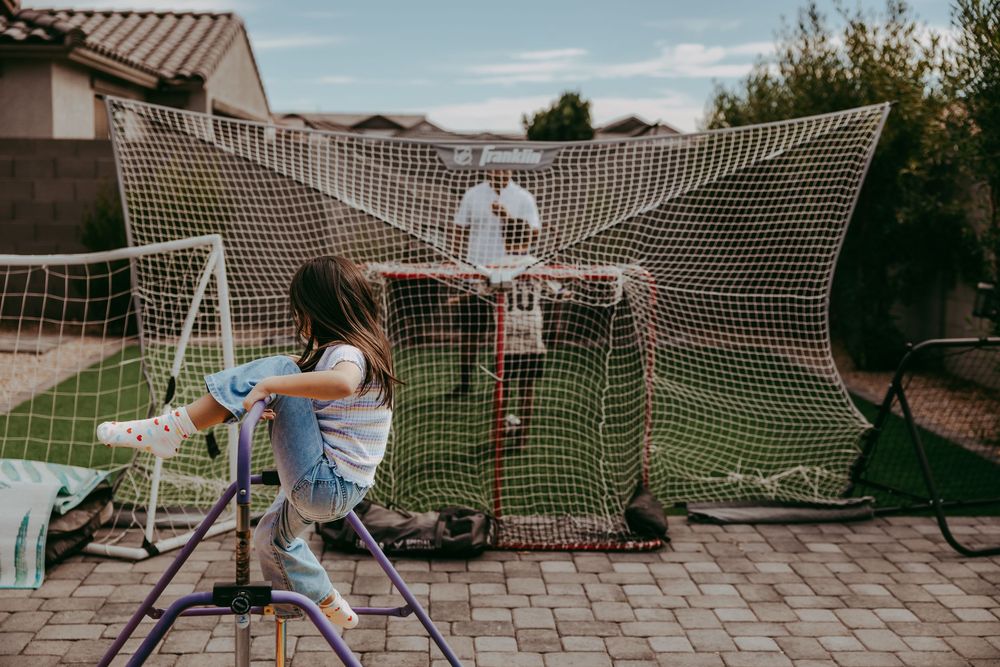 A girl plays on a gymnastic bar in a backyard while a person stands near a large sports net on a paved patio.