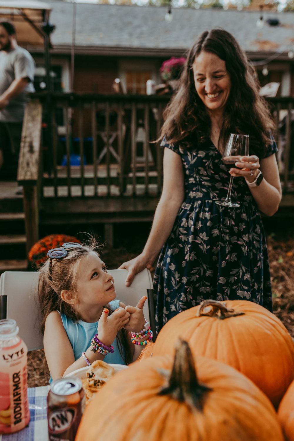 Woman in floral dress holding wine glass smiles at young girl near pumpkins at outdoor fall Halloween gathering.