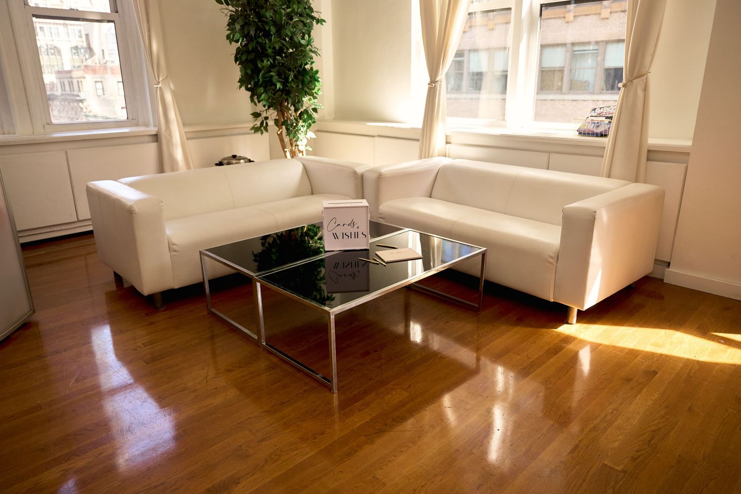 Modern white sectional sofa with glass coffee table on hardwood floors in bright office lounge with large windows.