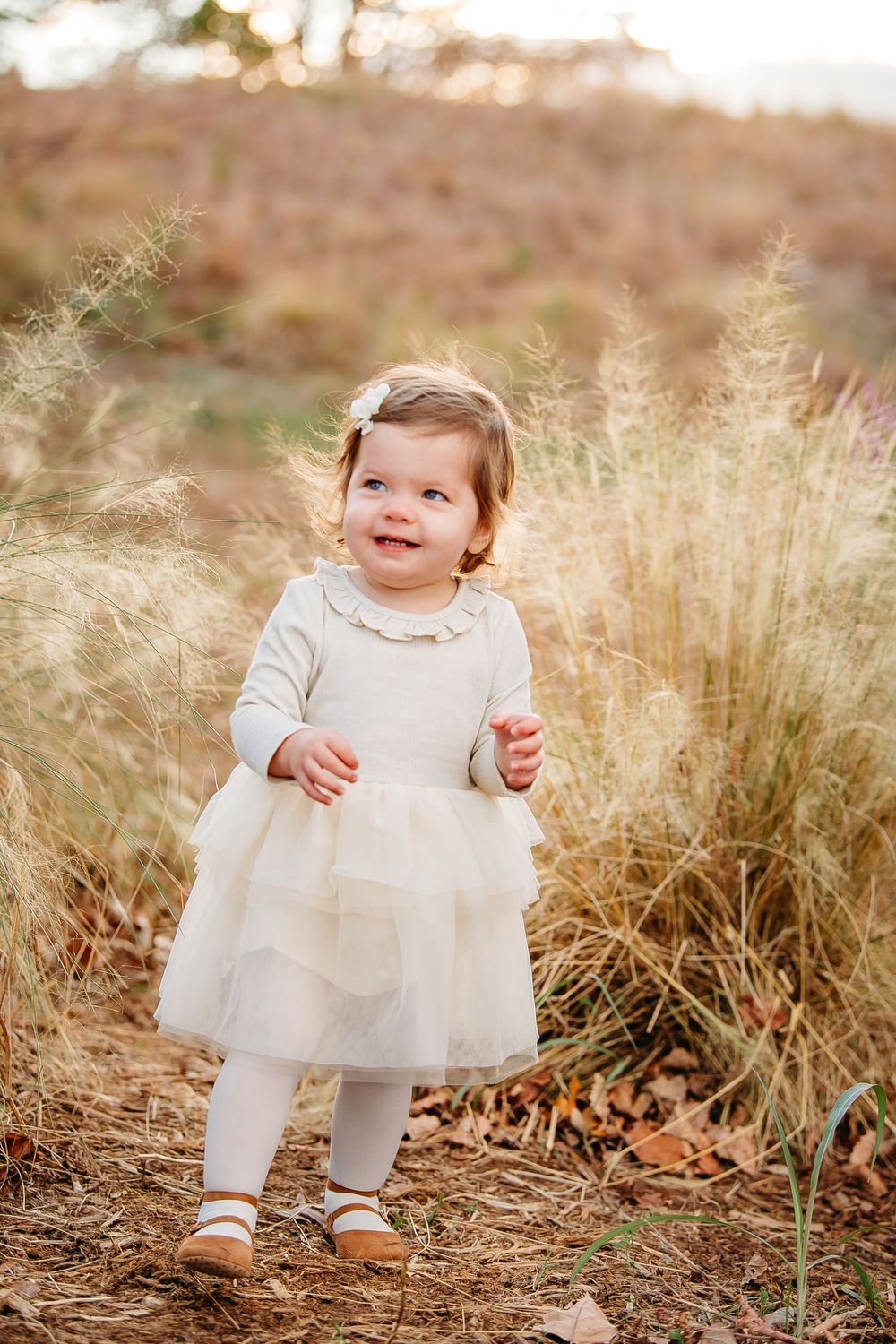 Adorable baby girl in white tutu dress smiling outdoors among tall golden grasses.
