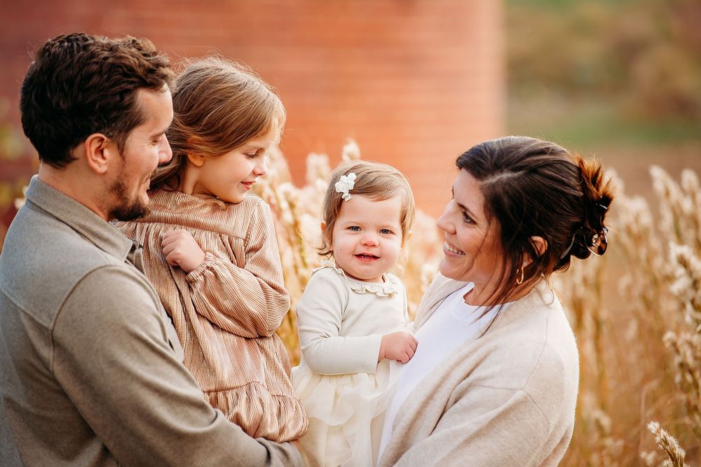 Loving parents holding two young daughters outdoors near golden wheat and brick wall.