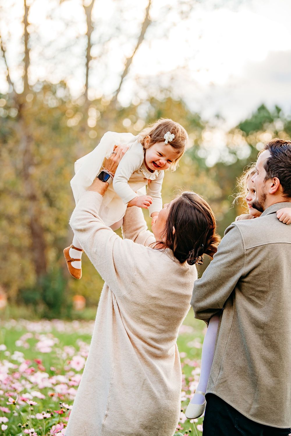 Parents joyfully lifting laughing toddler girl in white dress in a flower-filled park.