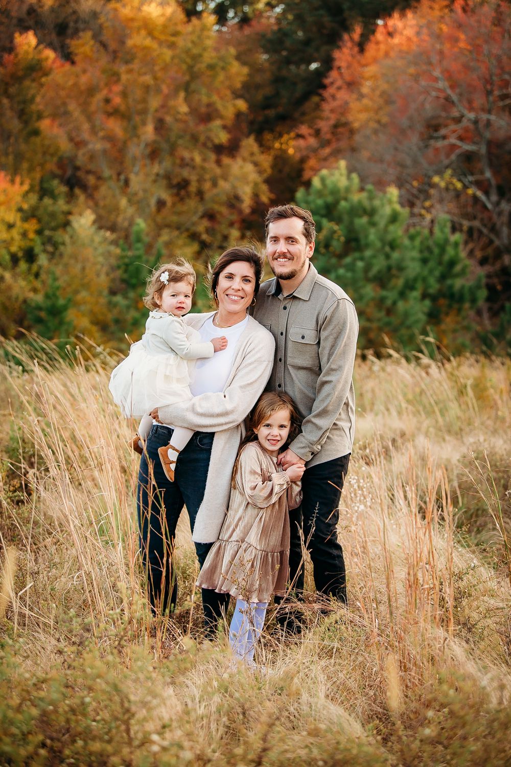 Happy family of four posing in tall golden grass with vibrant fall foliage behind them.