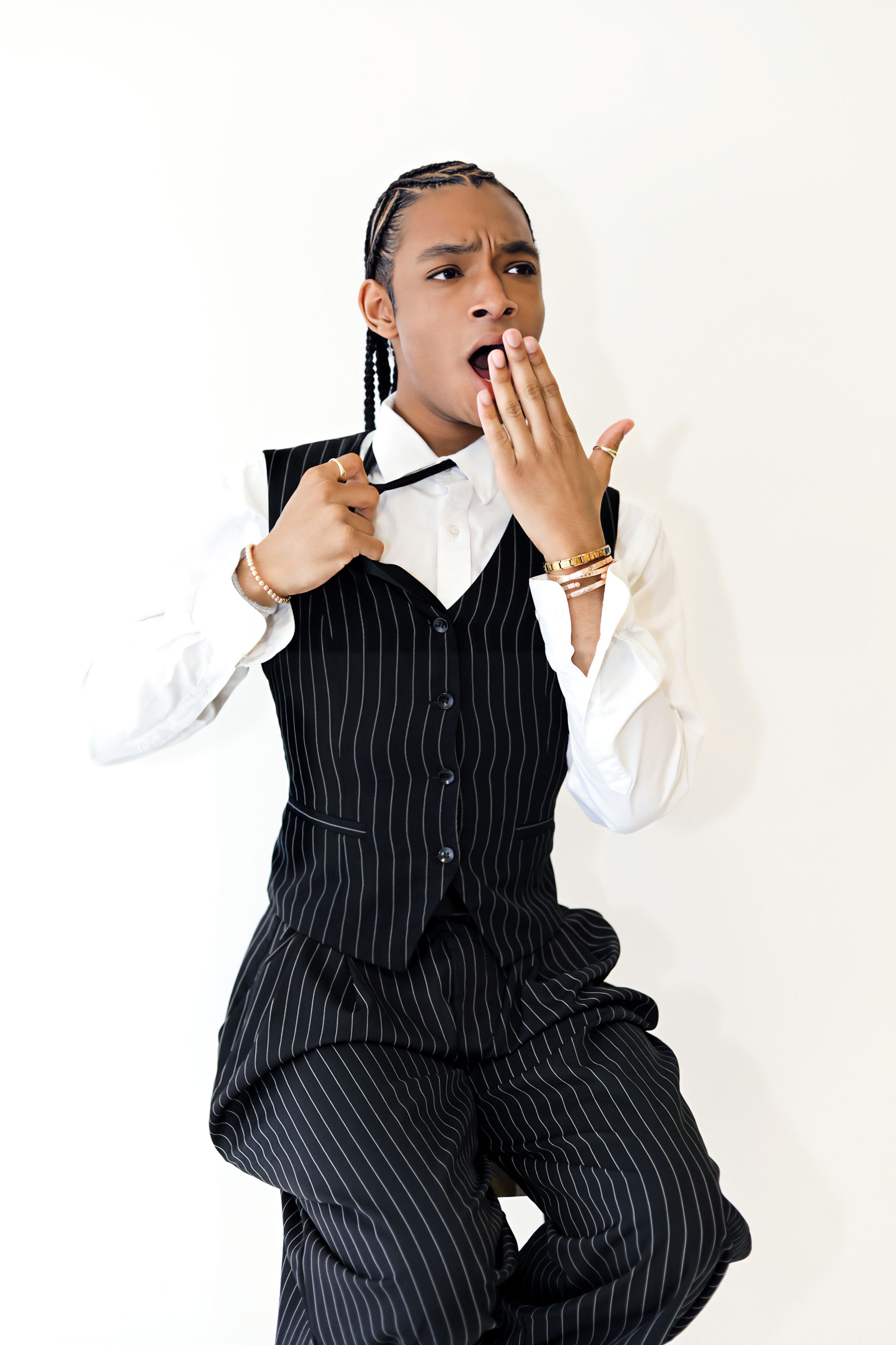 Young man in pinstripe vest and white shirt with braids, looking shocked with hand over mouth.
