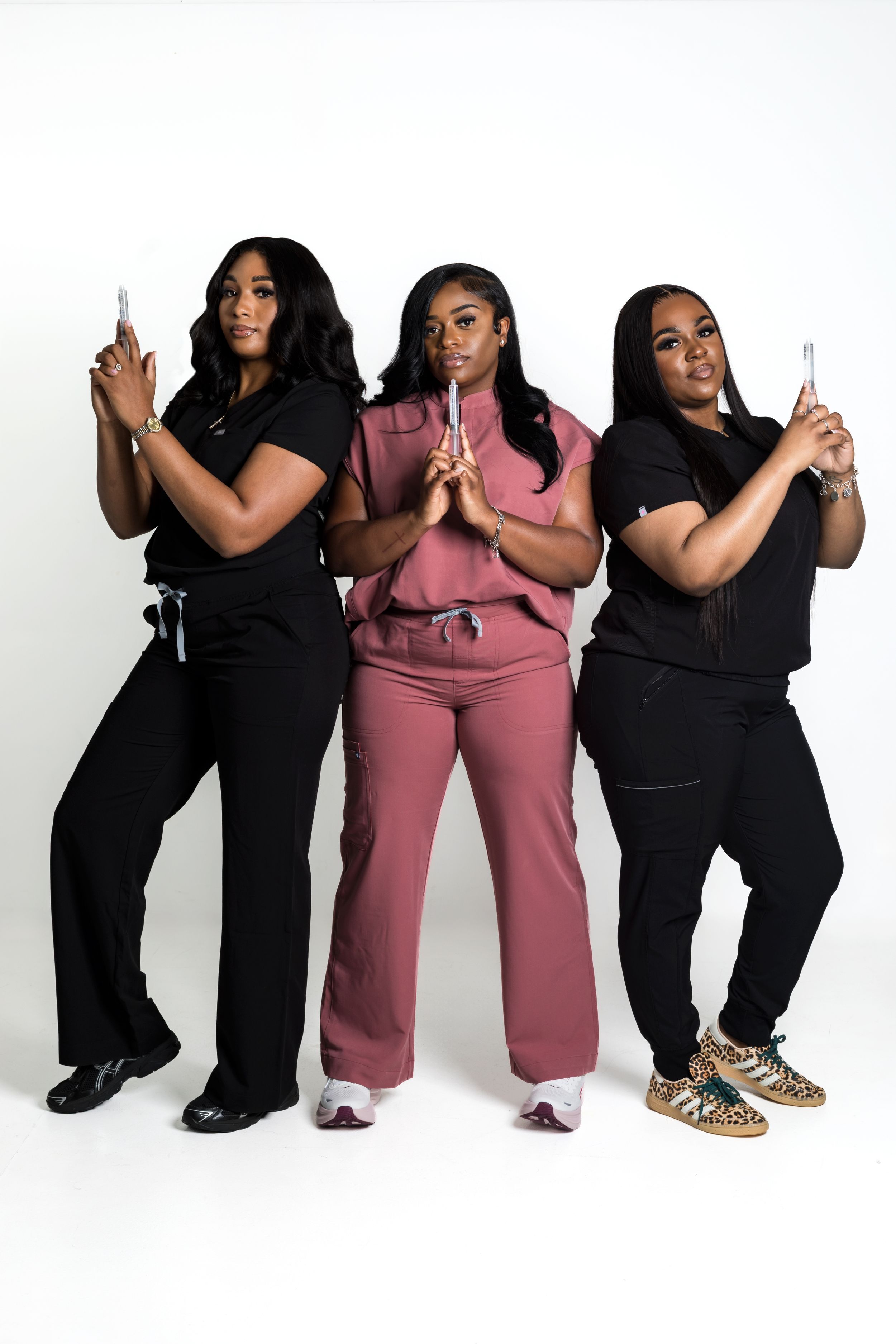 Three women in medical scrubs pose confidently holding syringes against a white background.