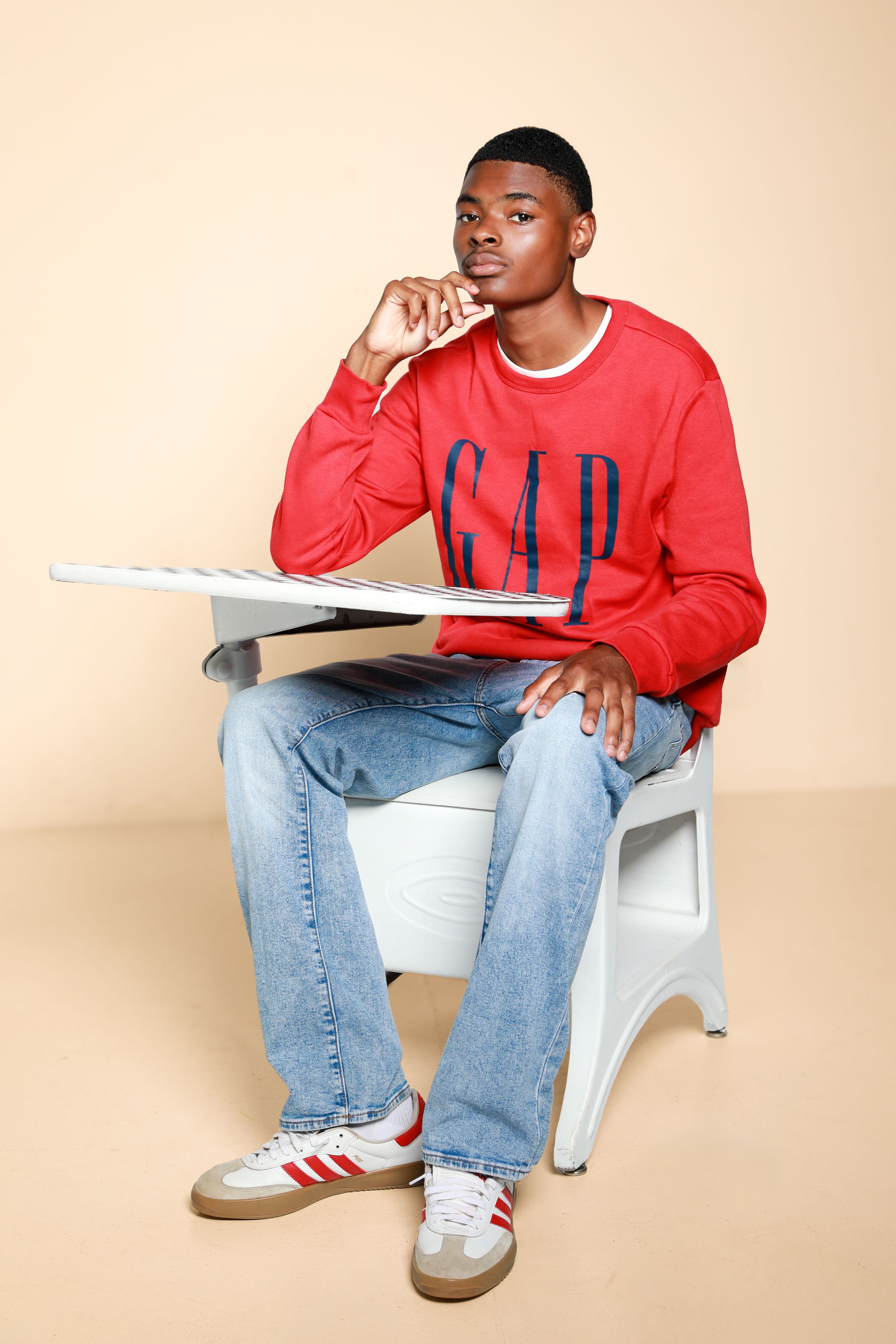 Young man in red GAP sweatshirt and jeans sits thoughtfully at a school desk wearing Adidas sneakers.