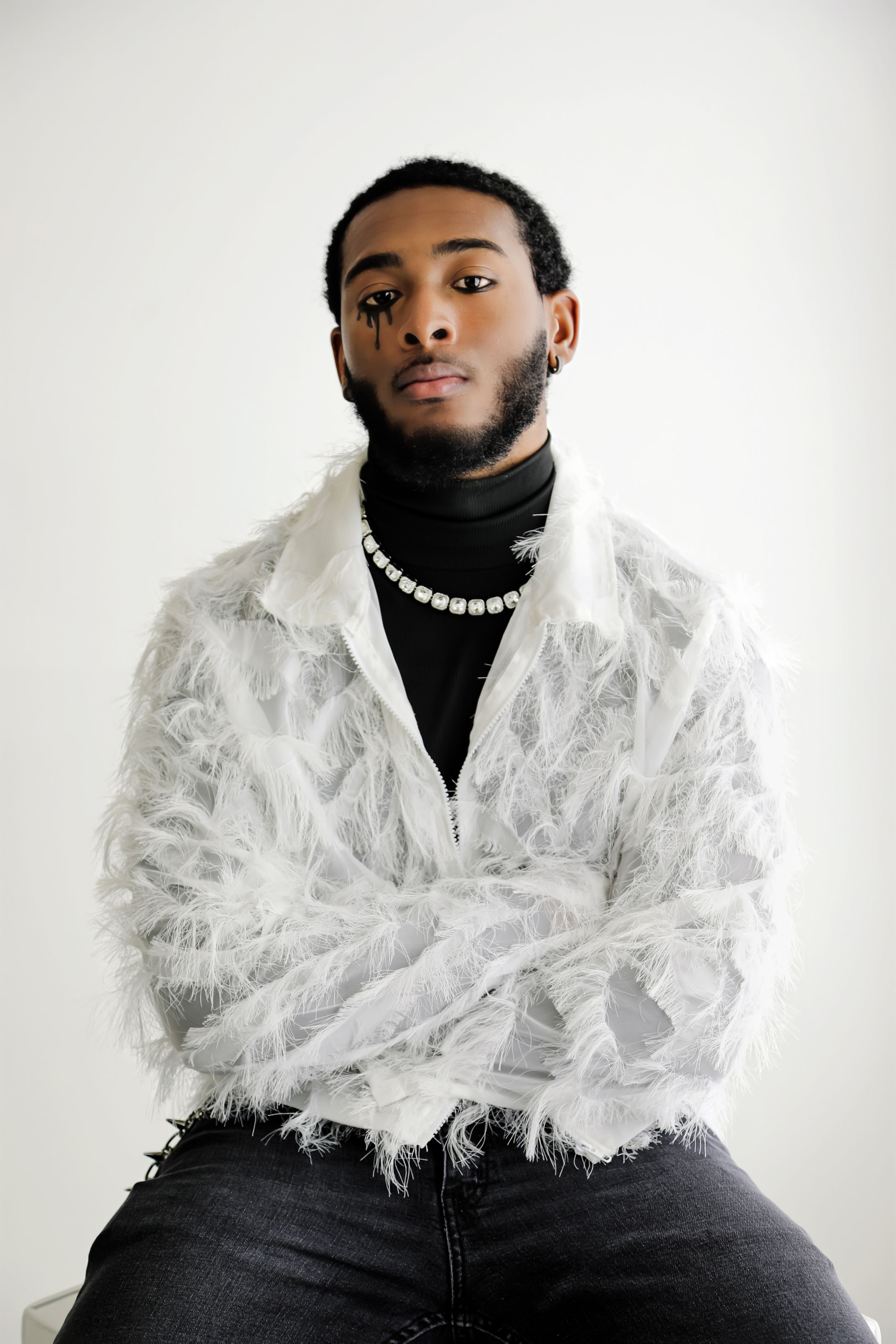 Young man in white feathered jacket, black turtleneck, pearl necklace, seated against white background.