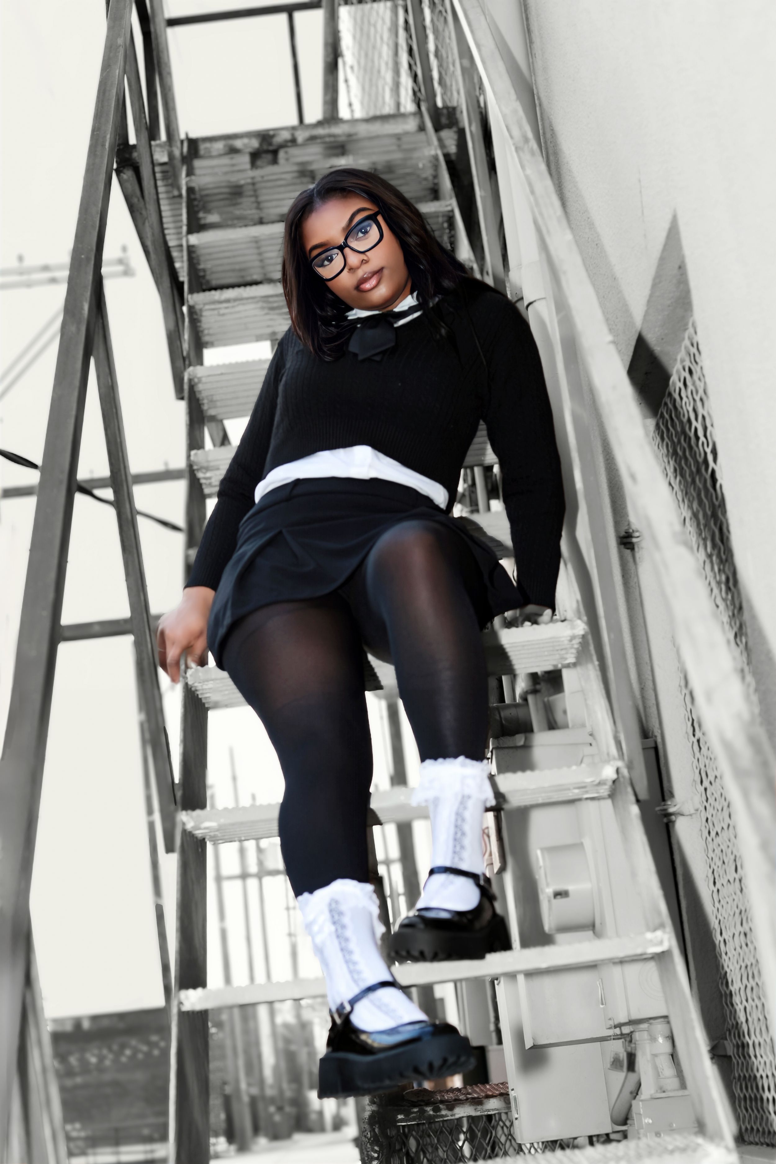 Young Black woman in glasses, black sweater, skirt, tights, and white socks poses on metal stairs outdoors.