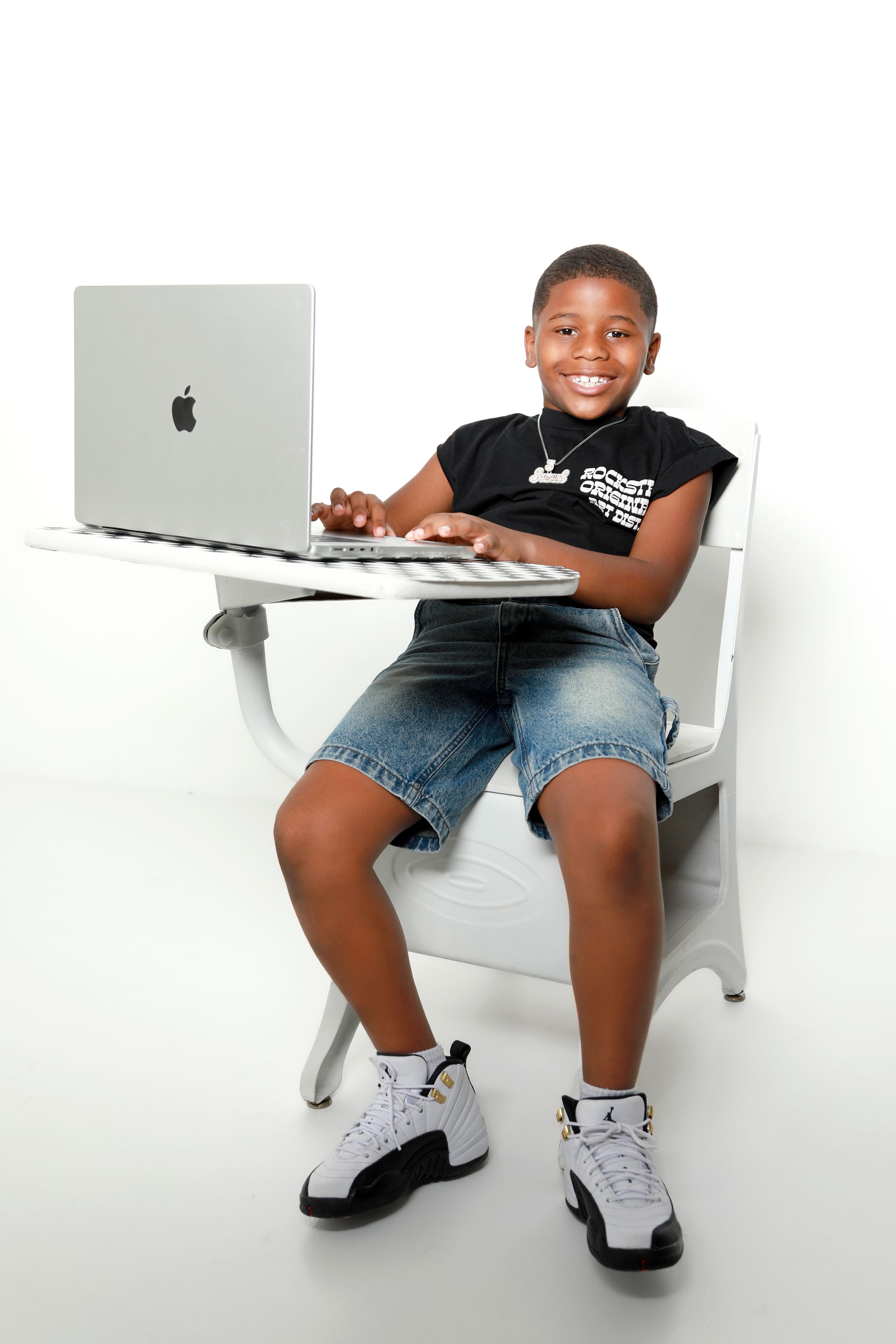 Smiling young boy sitting at a school desk with an Apple MacBook laptop, wearing jeans and Air Jordans.