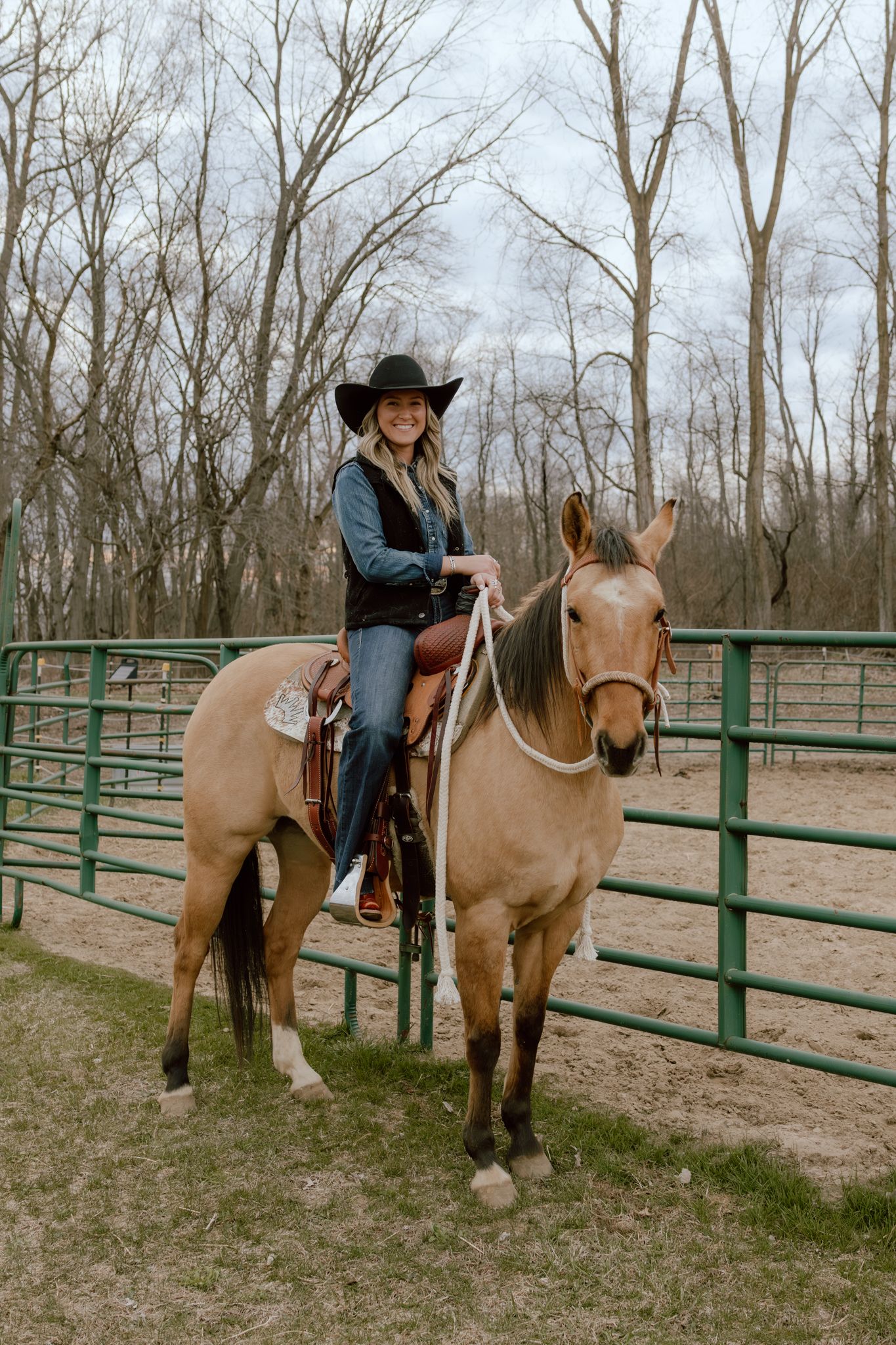 Smiling woman in black cowboy hat riding a buckskin horse in a green metal round pen with bare trees.