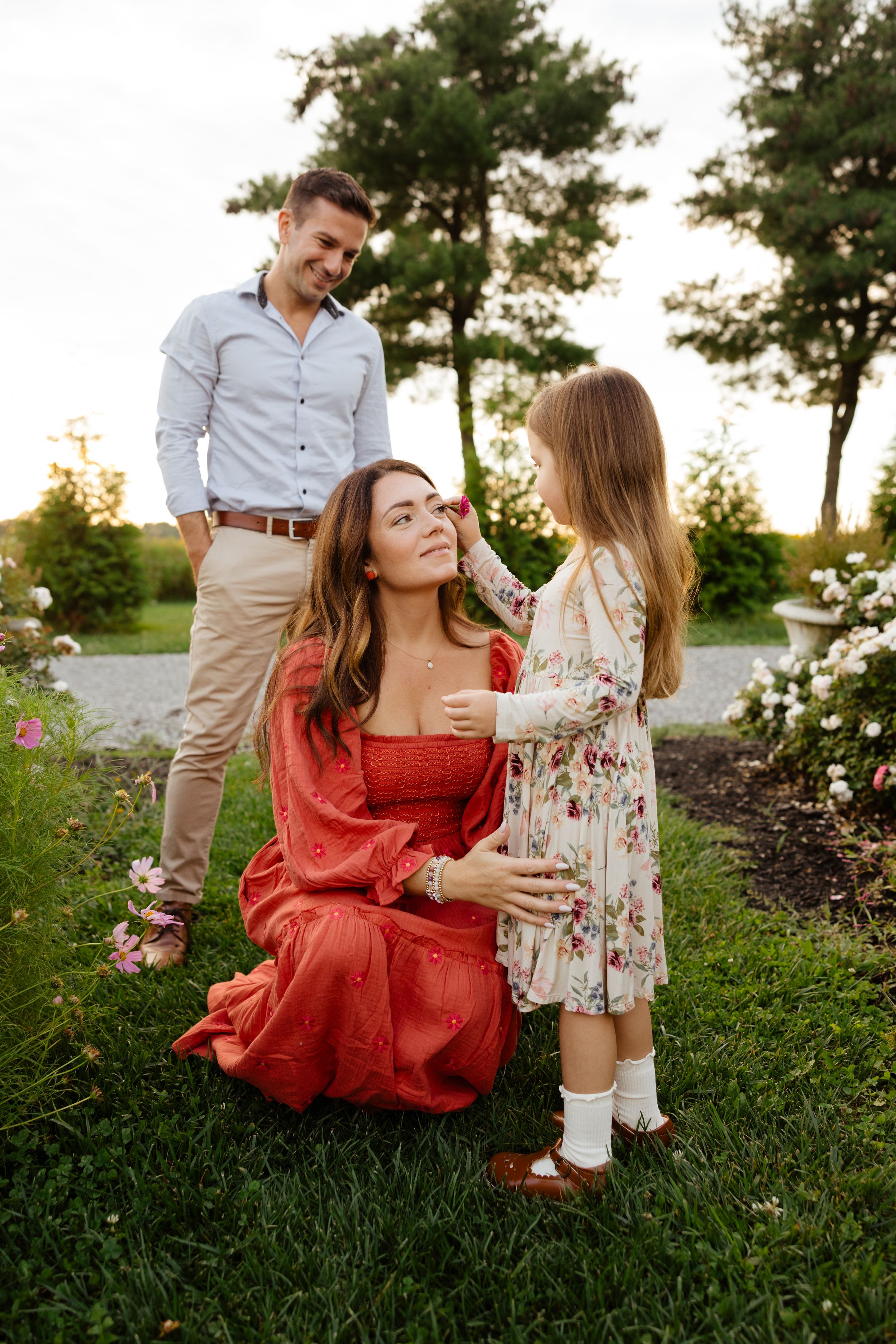 A family of three poses in a garden, with a little girl touching her mother's face while dad smiles warmly.