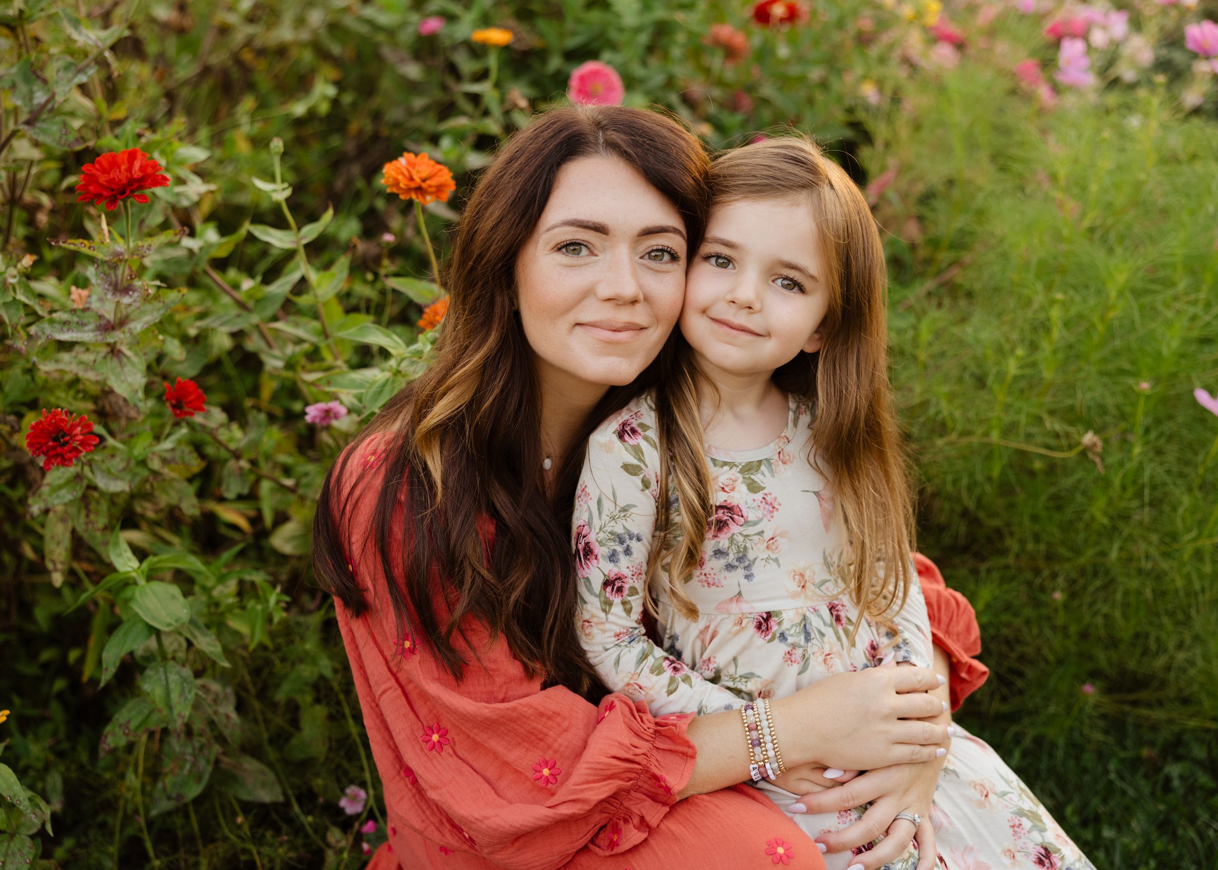 A woman and young girl smile together in a lush flower garden, both wearing floral dresses in a warm outdoor portrait.