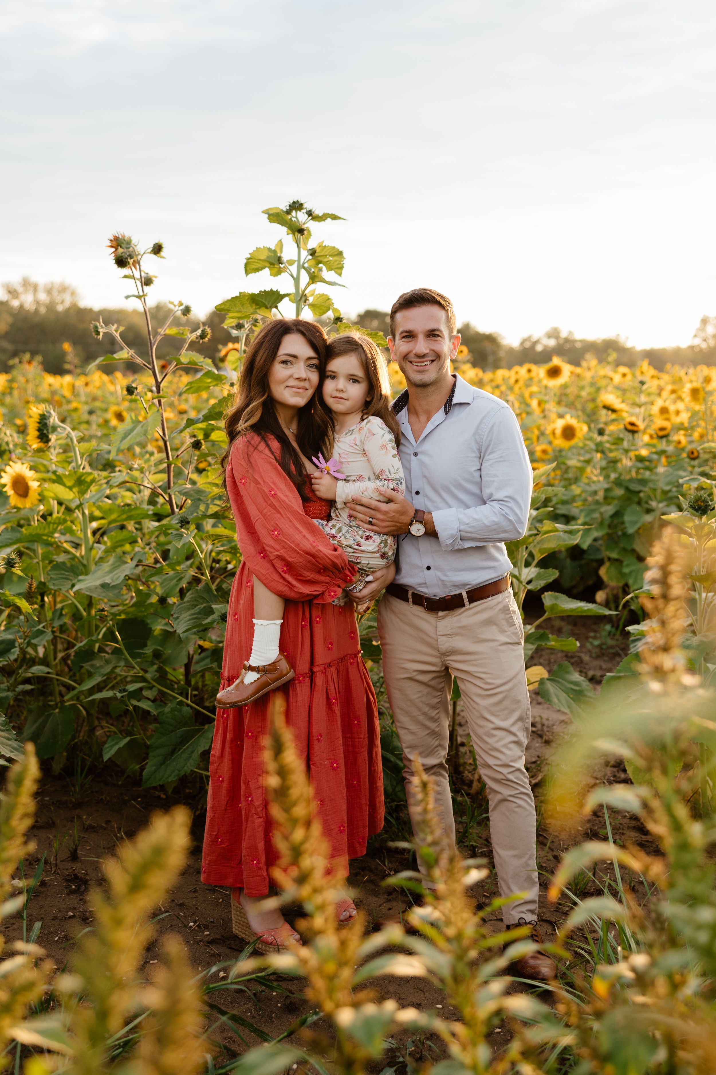 A smiling family of three poses in a vibrant sunflower field, with mom in red dress holding their toddler.