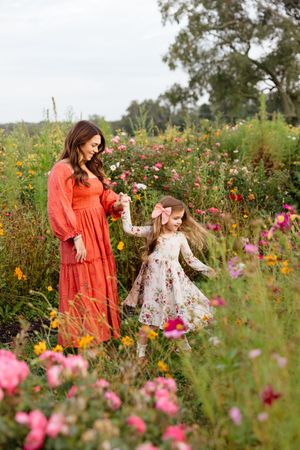 Mother in red dress holds daughter's hand in a colorful wildflower field, both wearing floral dresses.