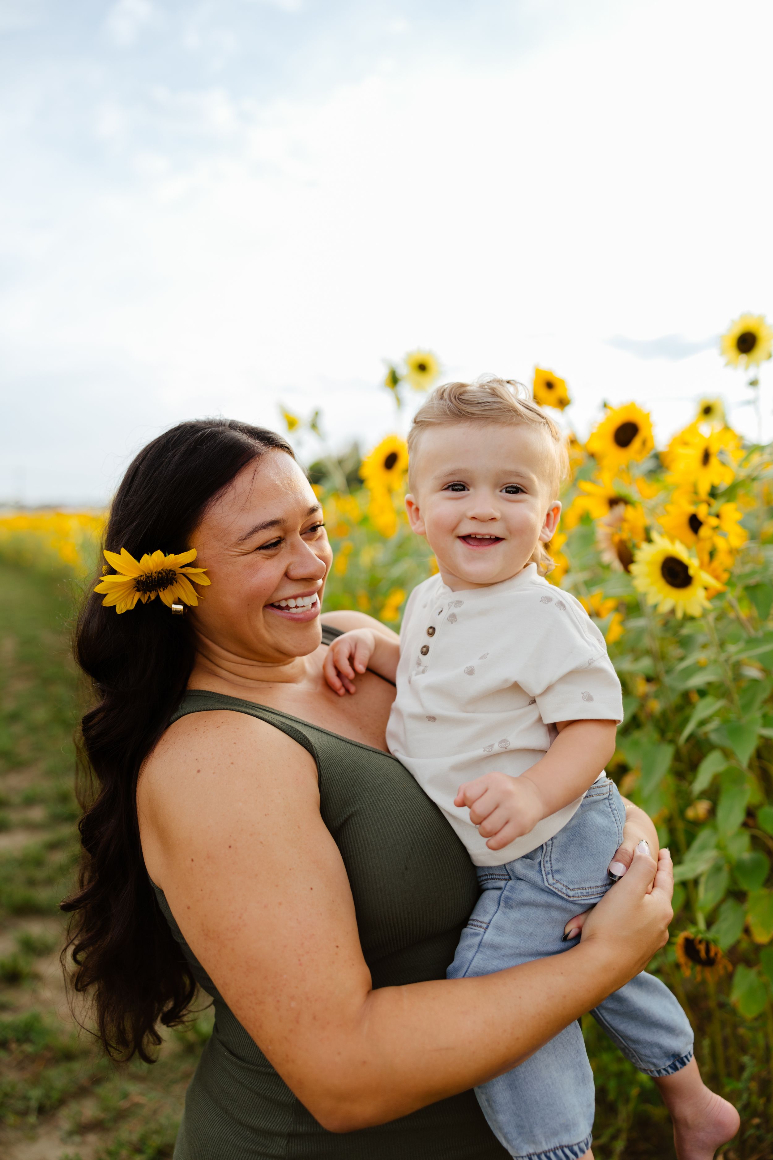 A joyful mother with a sunflower in her hair holds her smiling toddler in a vibrant sunflower field.