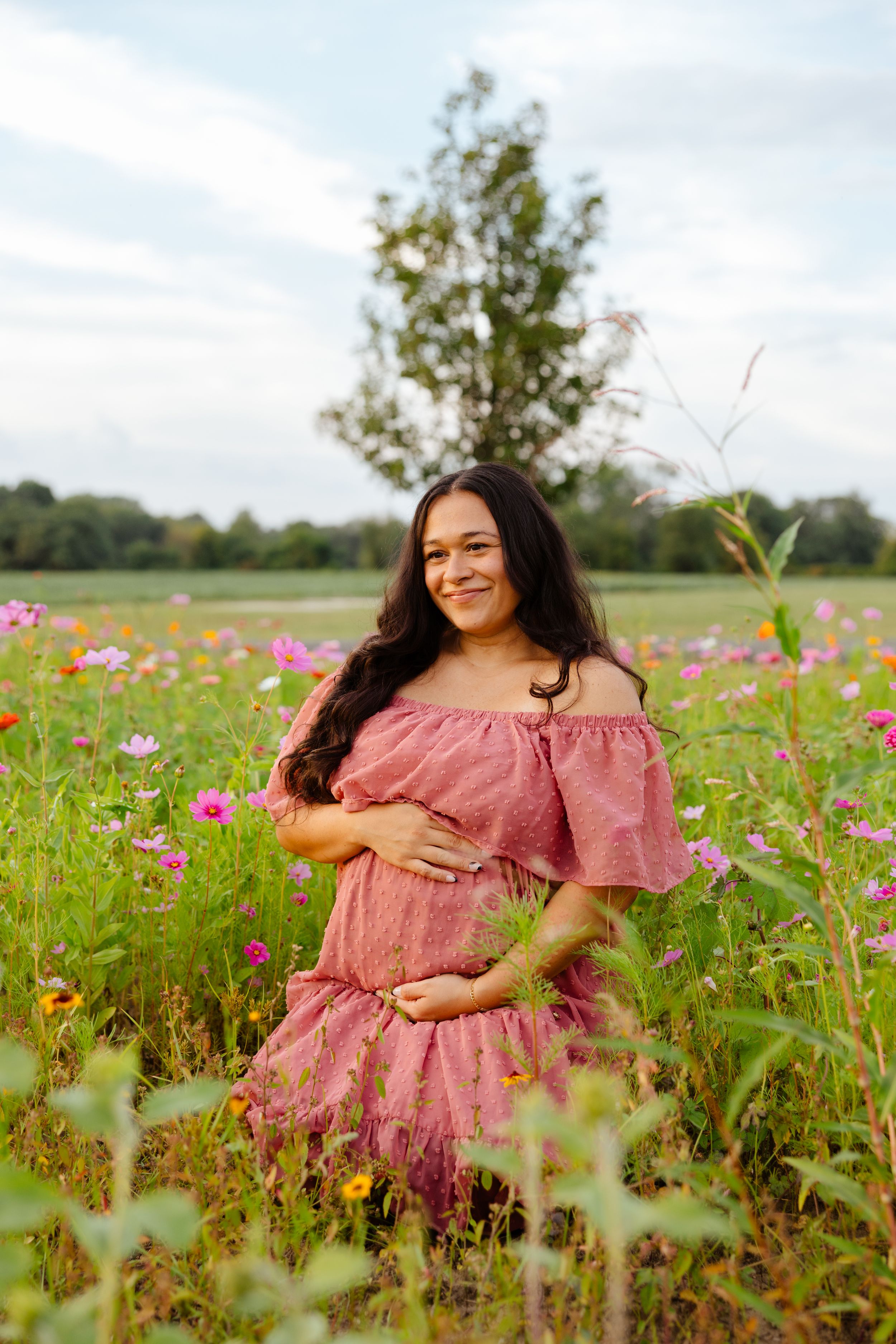 Smiling pregnant woman in pink off-shoulder dress standing in colorful wildflower field with trees and cloudy sky backdrop.