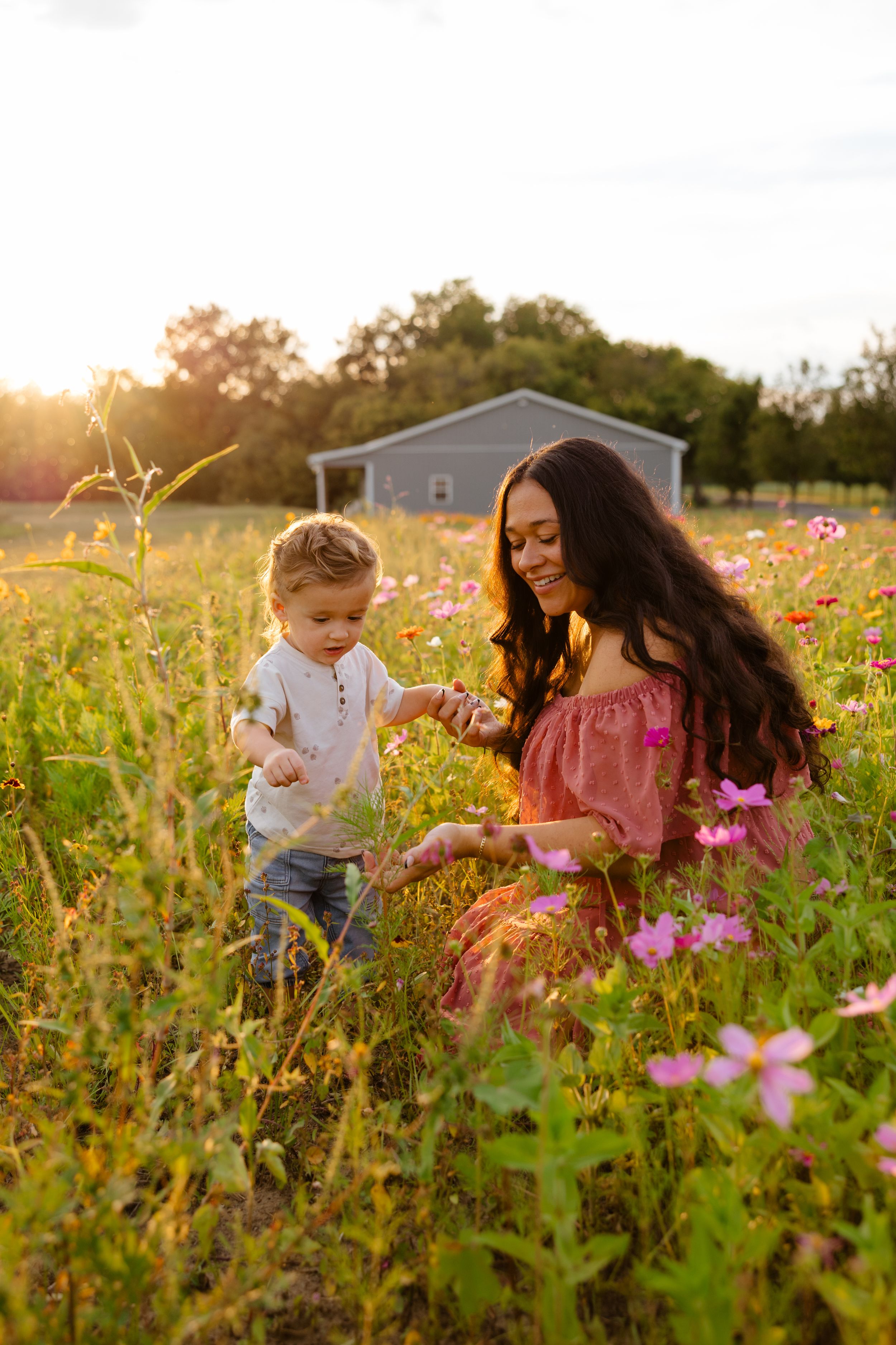Mother and toddler exploring a wildflower field at golden hour with a farmhouse in the background.