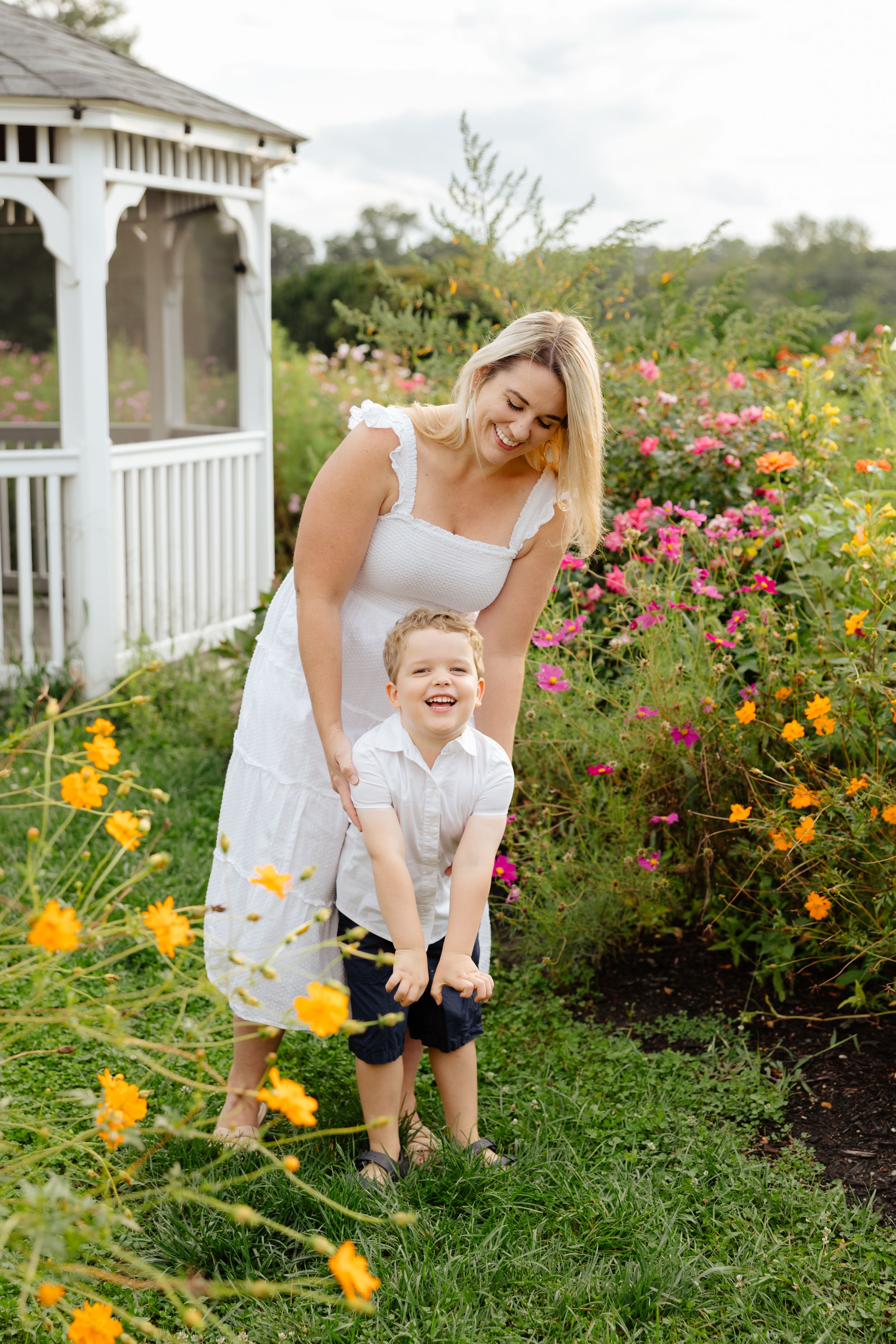 A smiling blonde woman in a white dress plays with a laughing young boy in a colorful garden near a white gazebo.