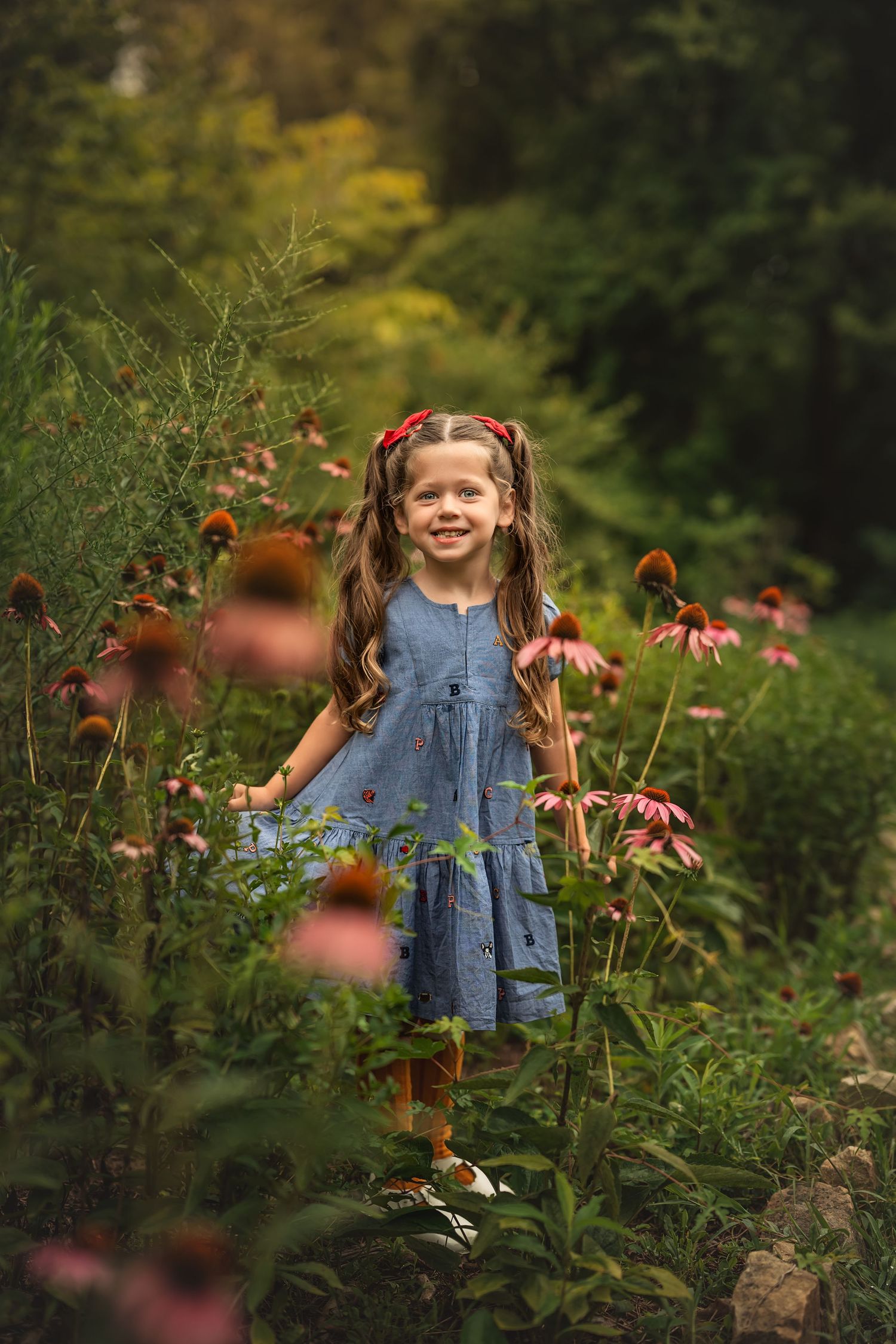 Young girl in blue dress smiling among pink echinacea flowers in a lush green garden.