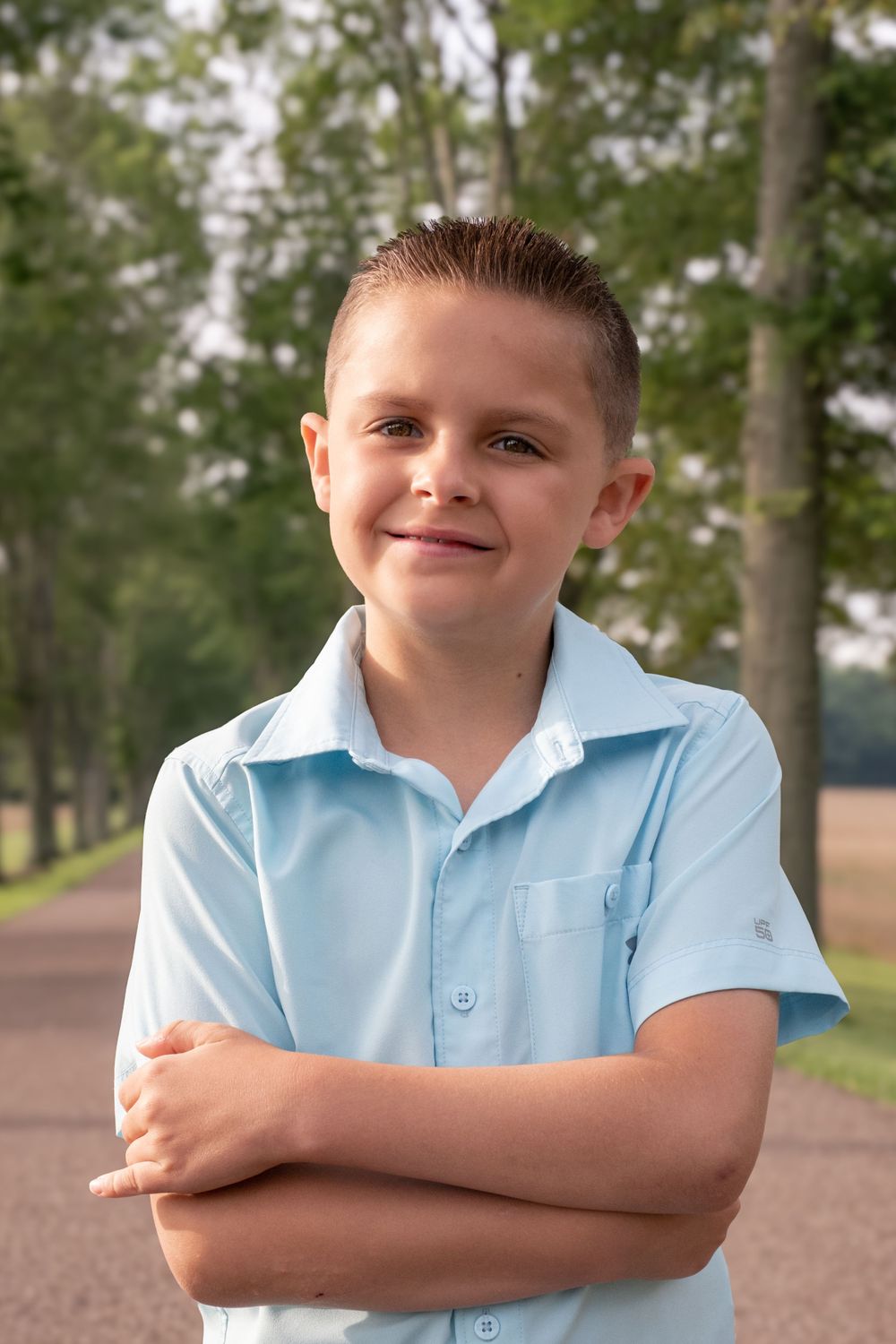 Smiling young boy in a light blue shirt standing with arms crossed on a tree-lined path outdoors. Uniontown, PA