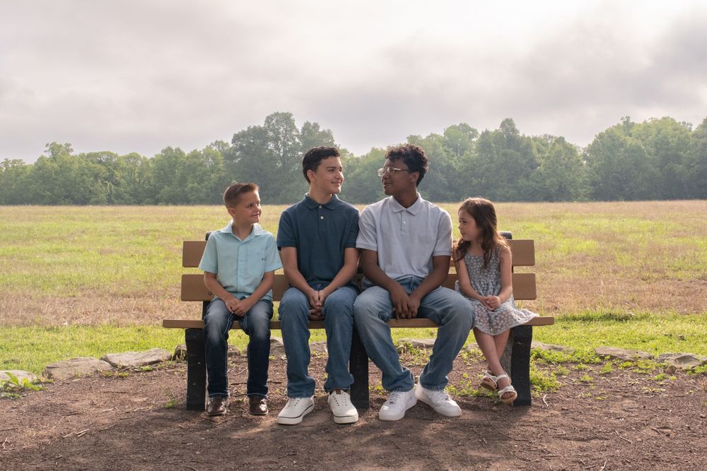 Four kids sitting on a wooden bench in a grassy field, smiling and chatting on a cloudy day. Uniontown, PA