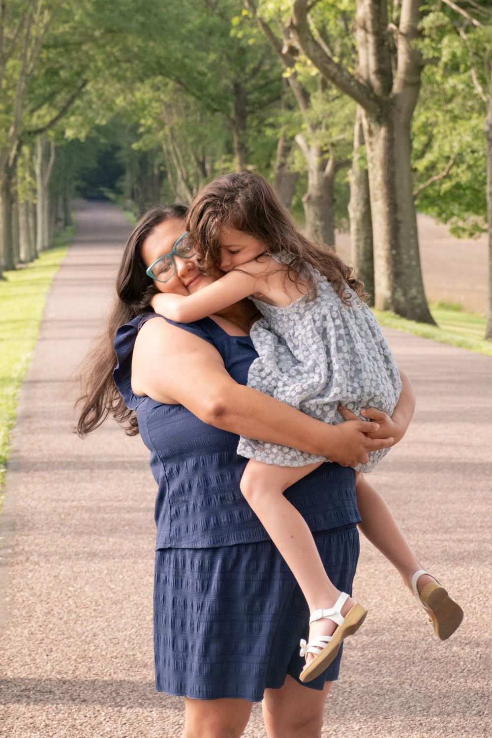 A mother in a navy dress hugs her young daughter on a tree-lined path, sharing a joyful embrace outdoors. Uniontown, PA