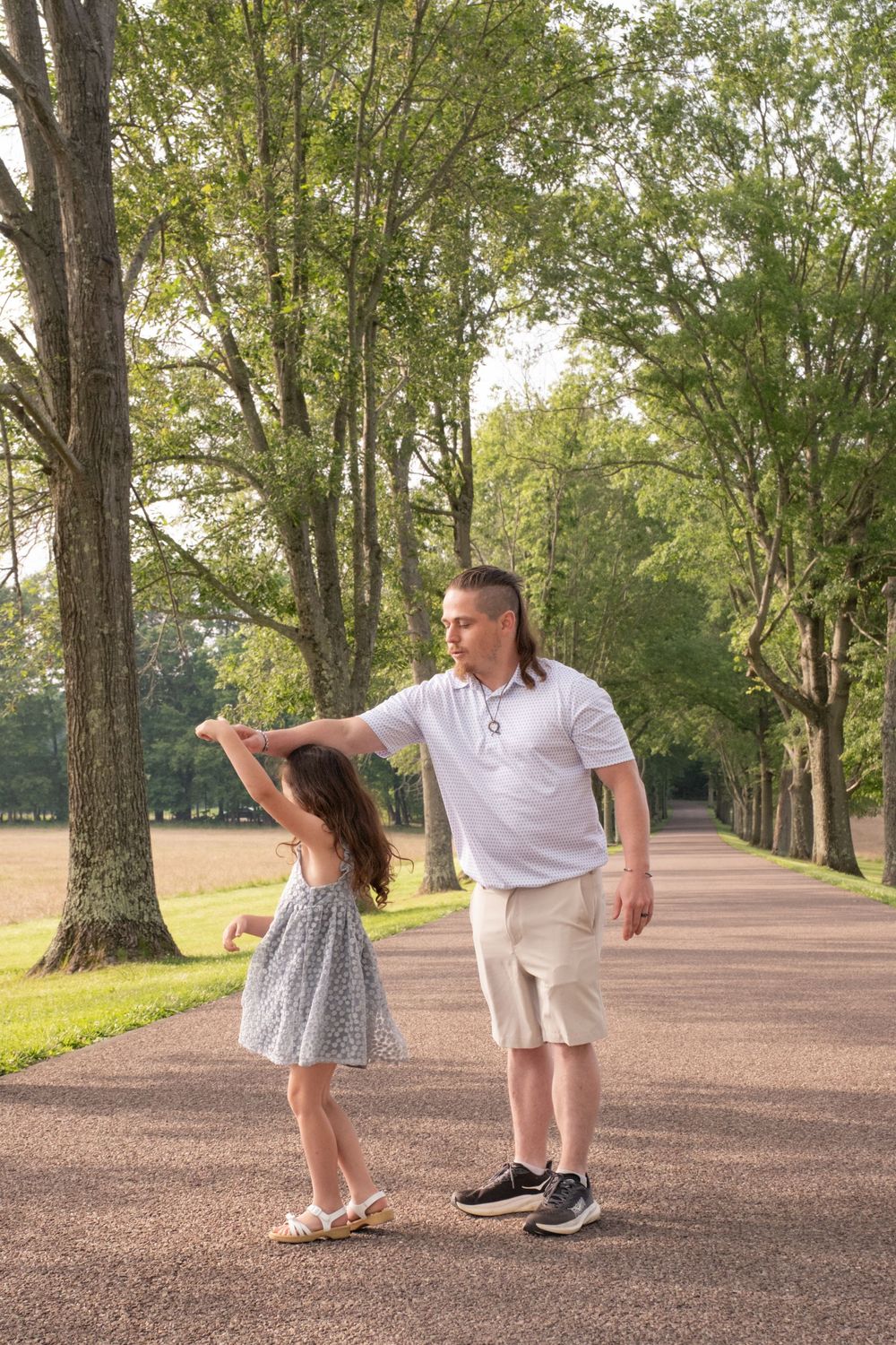 A father and daughter dancing together on a tree-lined path in a park on a sunny day. Uniontown, PA