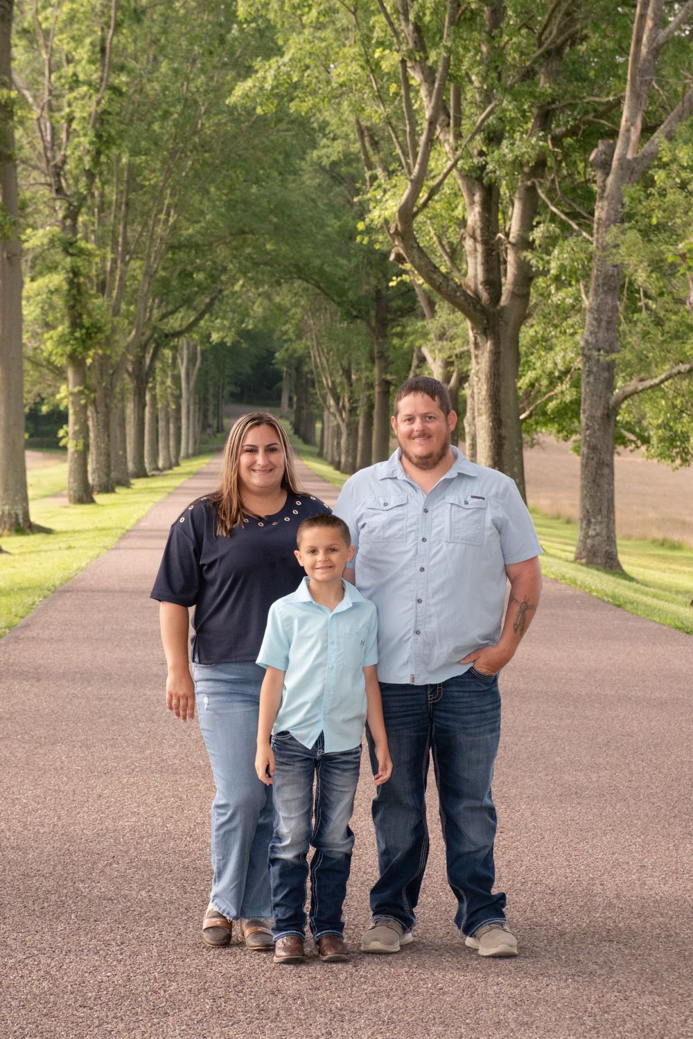 Family of three posing on a tree-lined path, parents flanking a young boy, all wearing jeans and casual shirts outdoors. Uniontown, PA