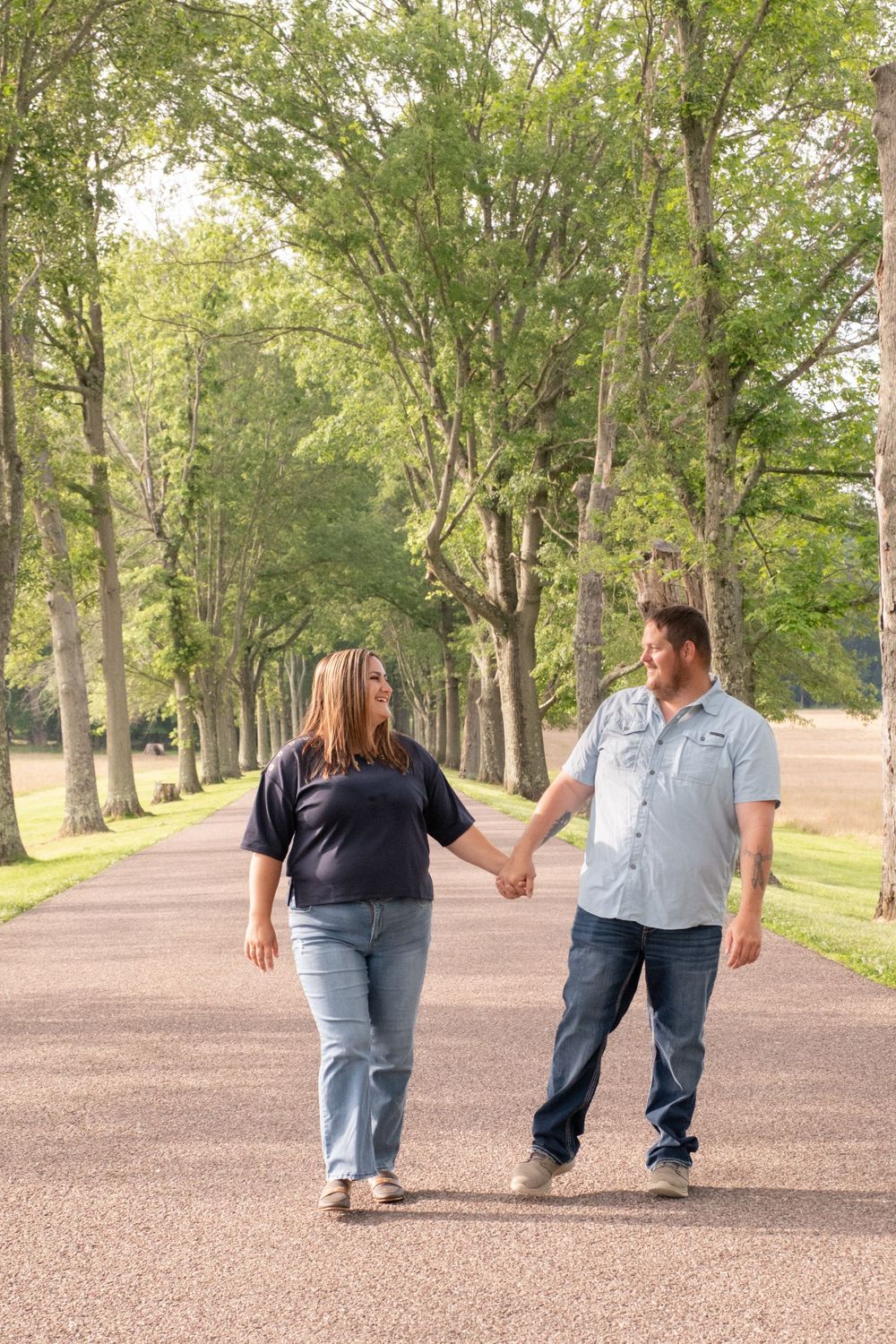 Couple holding hands walking down a tree-lined path in a park, wearing casual jeans and button-up shirts. Uniontown, PA