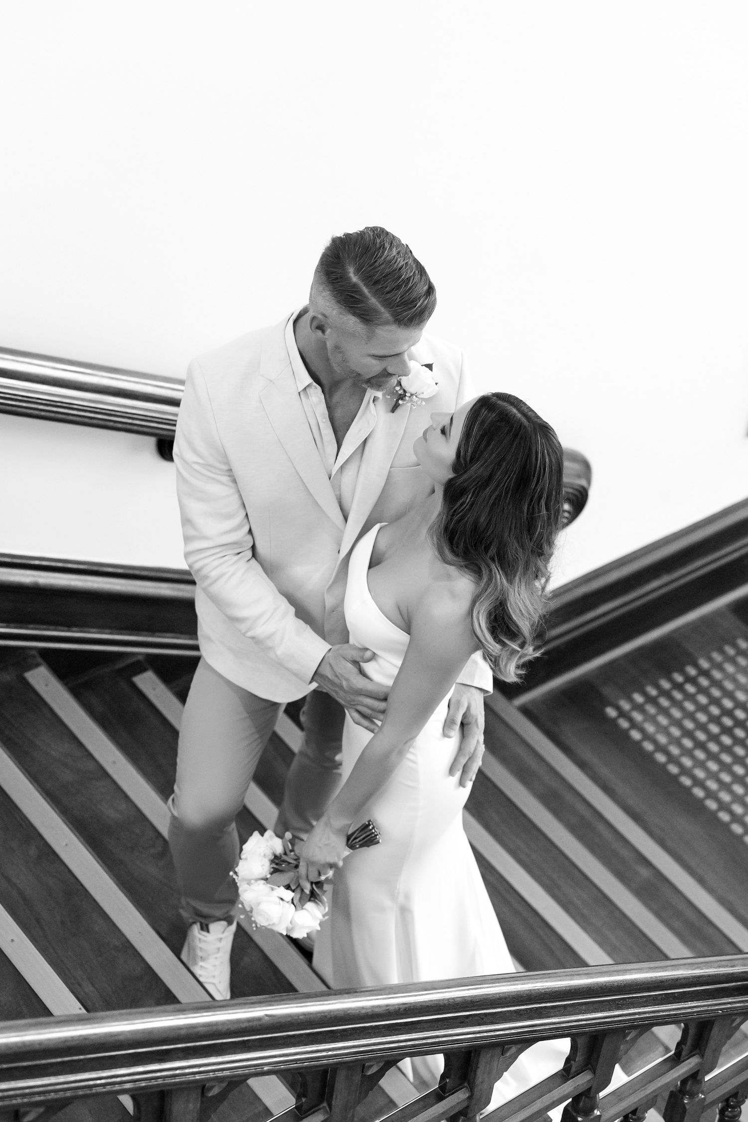 Black and white photo of a couple kissing on wooden stairs, bride holding bouquet in elegant wedding attire.