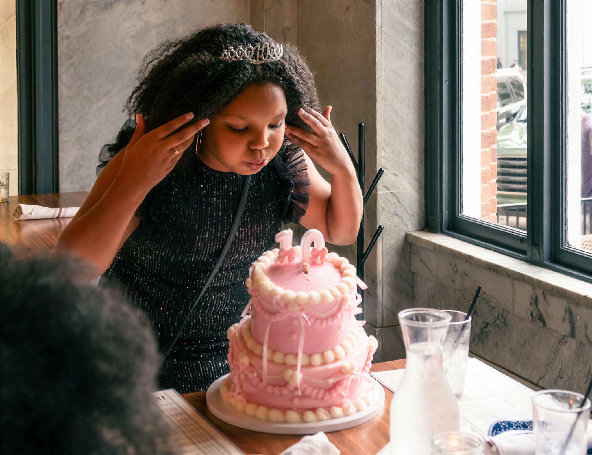 A young girl sits at a restaurant table with a two-tier pink birthday cake with the number 10, holding her head in her hands.