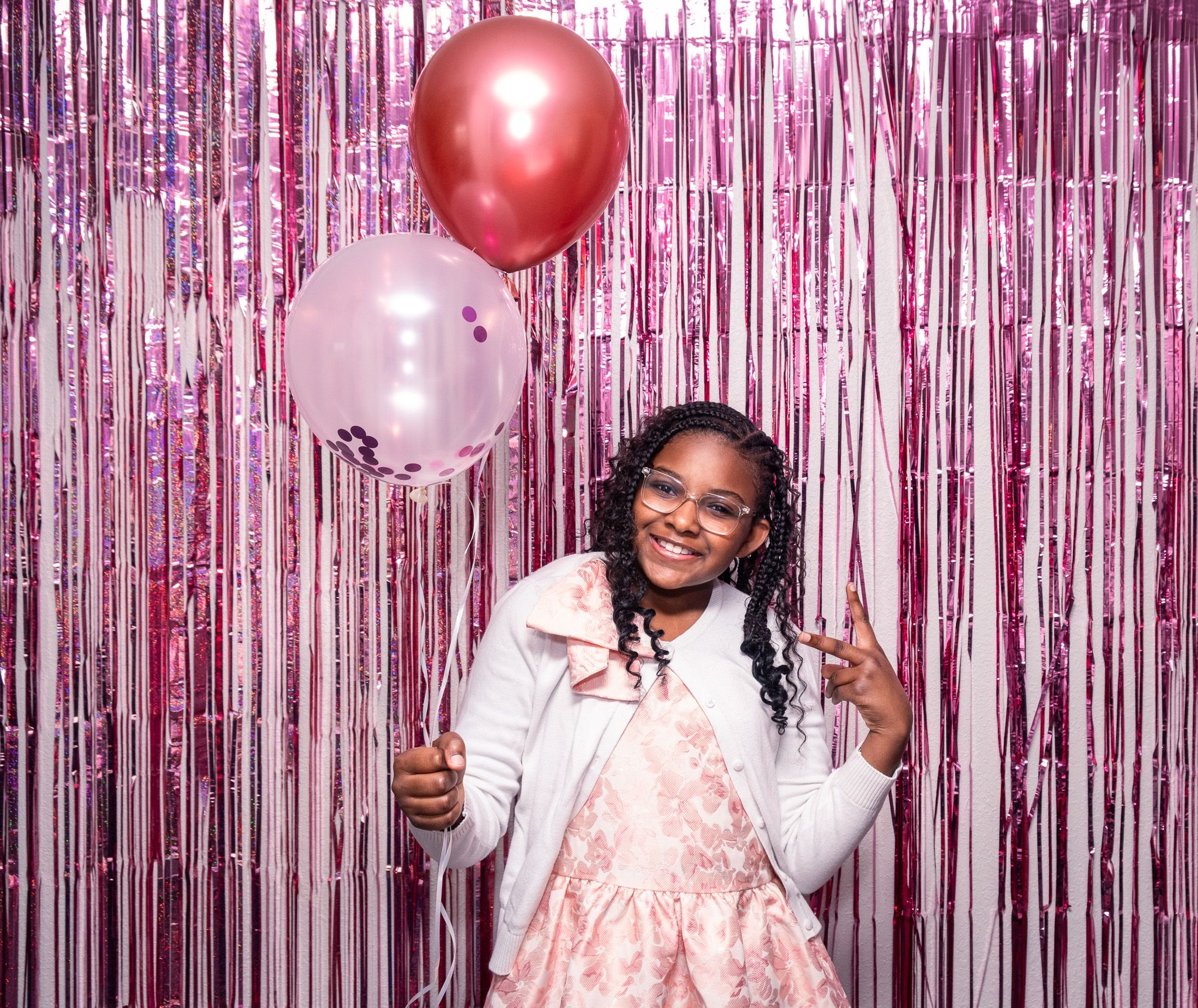 Young girl in pink dress holding balloons in front of pink tinsel fringe backdrop at a birthday party.