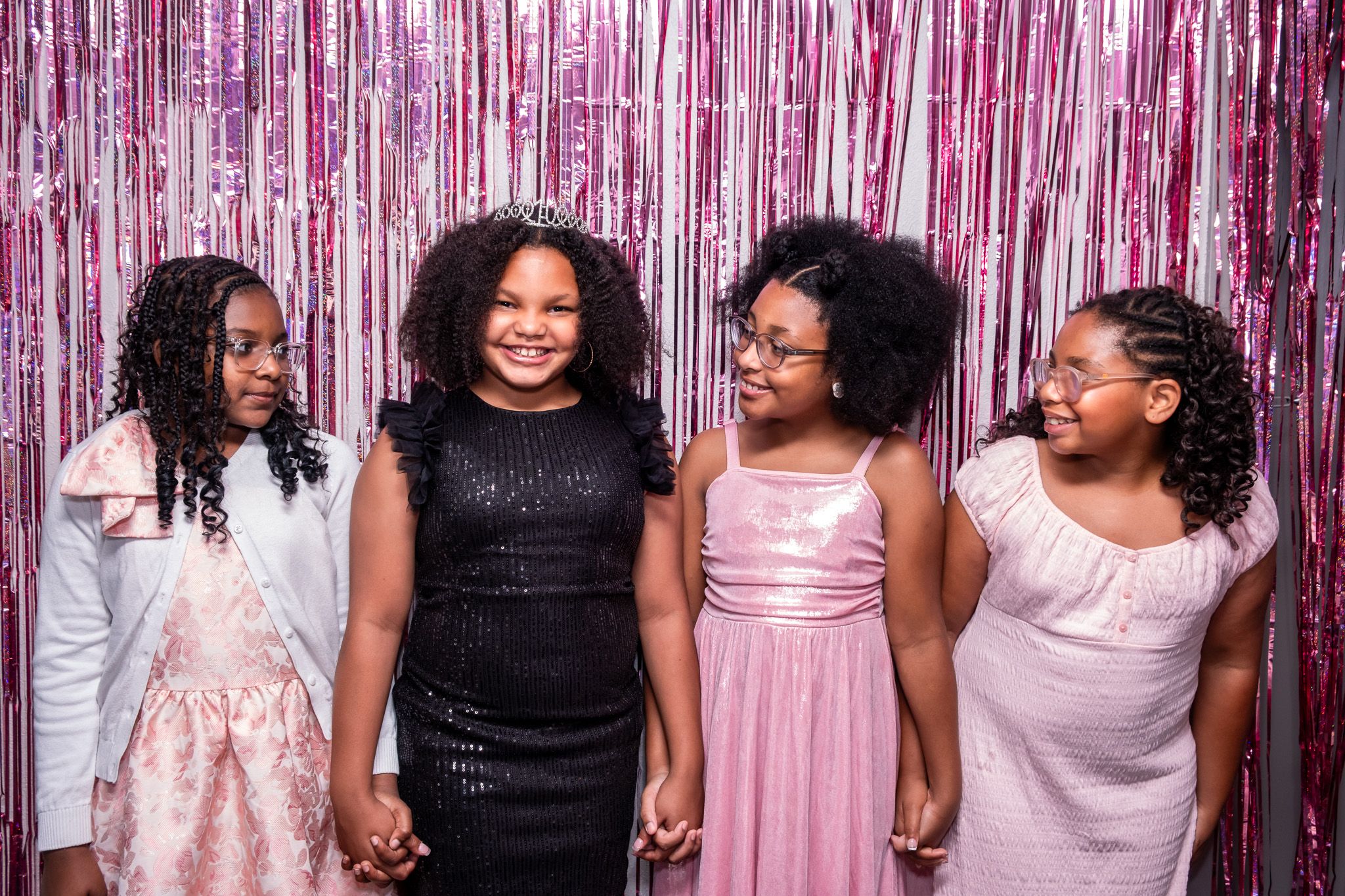 Four girls in formal dresses smile and hold hands in front of a pink metallic fringe backdrop.