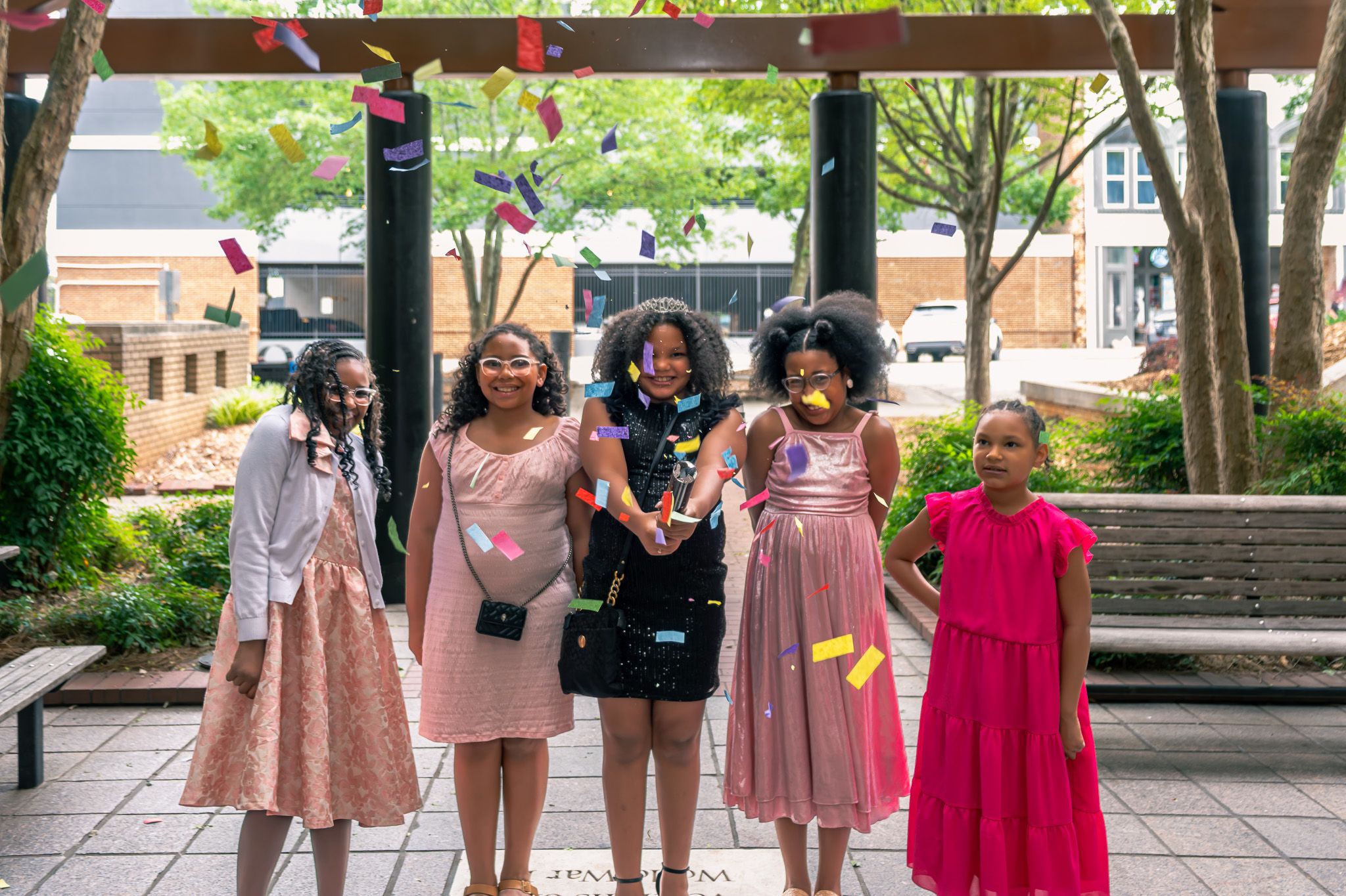 Five young girls in colorful dresses pose together outdoors at a festive event with trees and confetti.