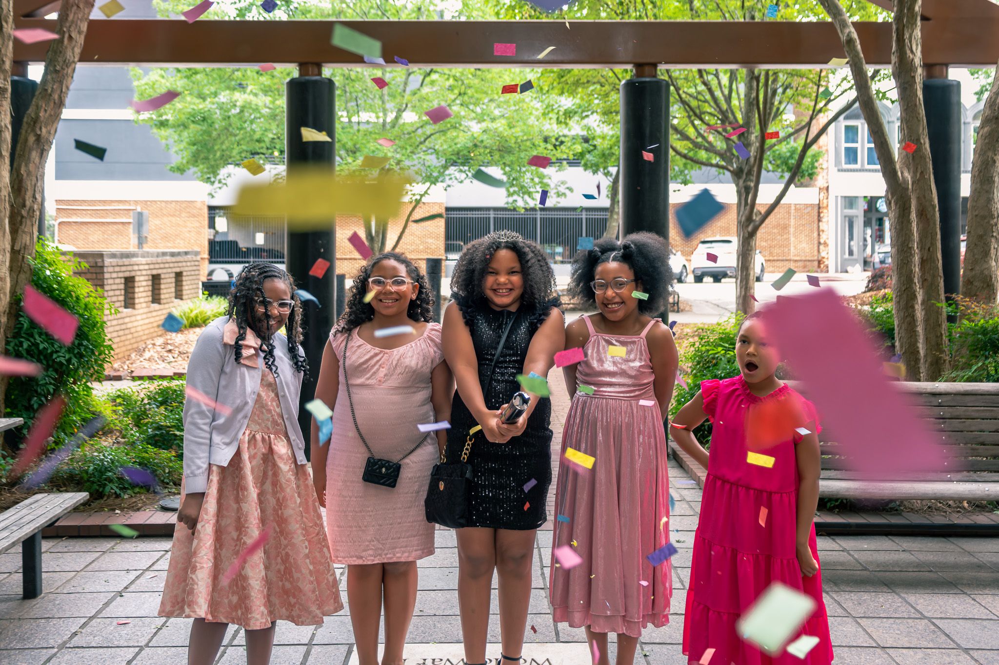 Five girls in colorful formal dresses celebrate outdoors with confetti falling around them.