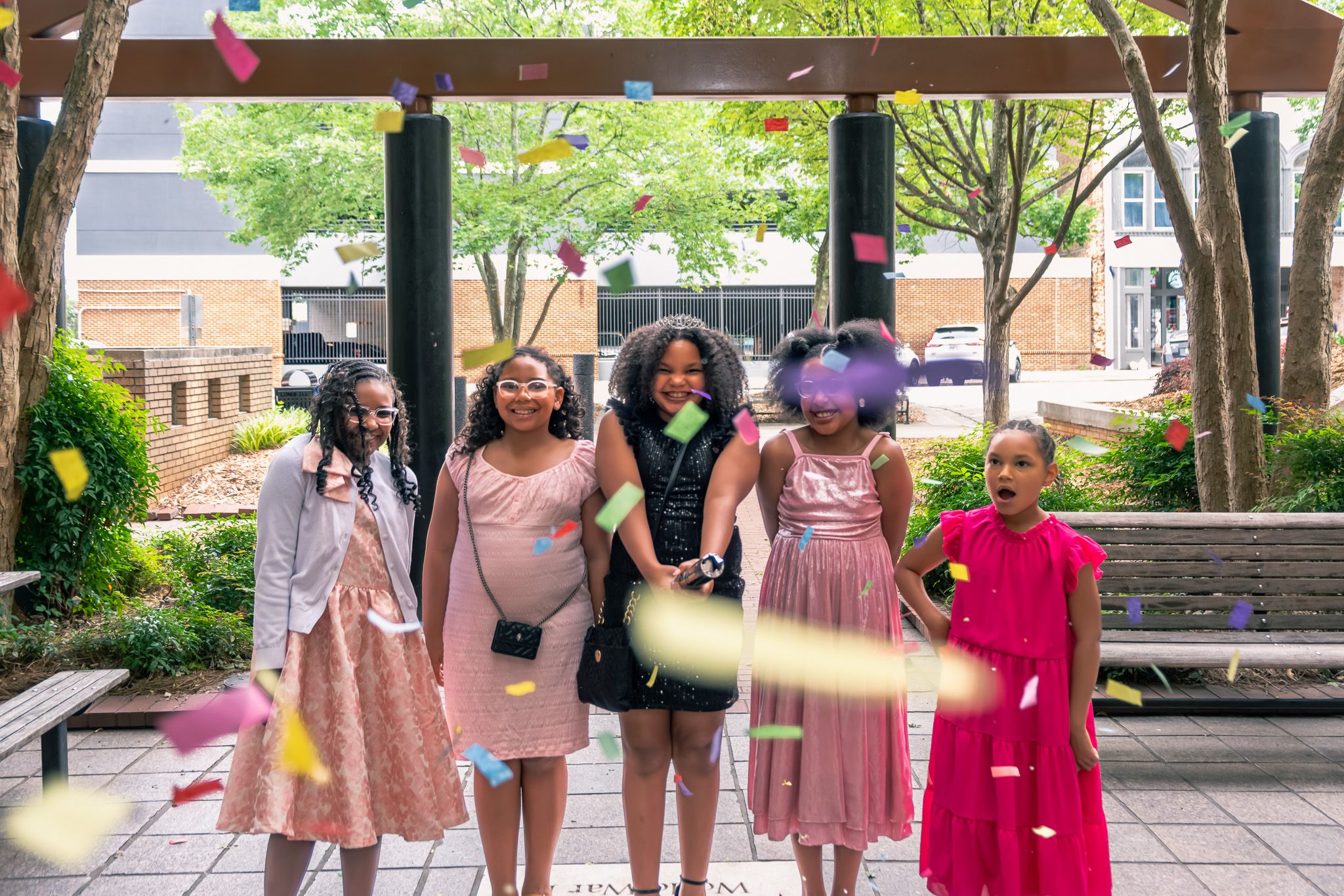 Five young girls in colorful dresses smile outdoors as confetti flies around them during a celebration.