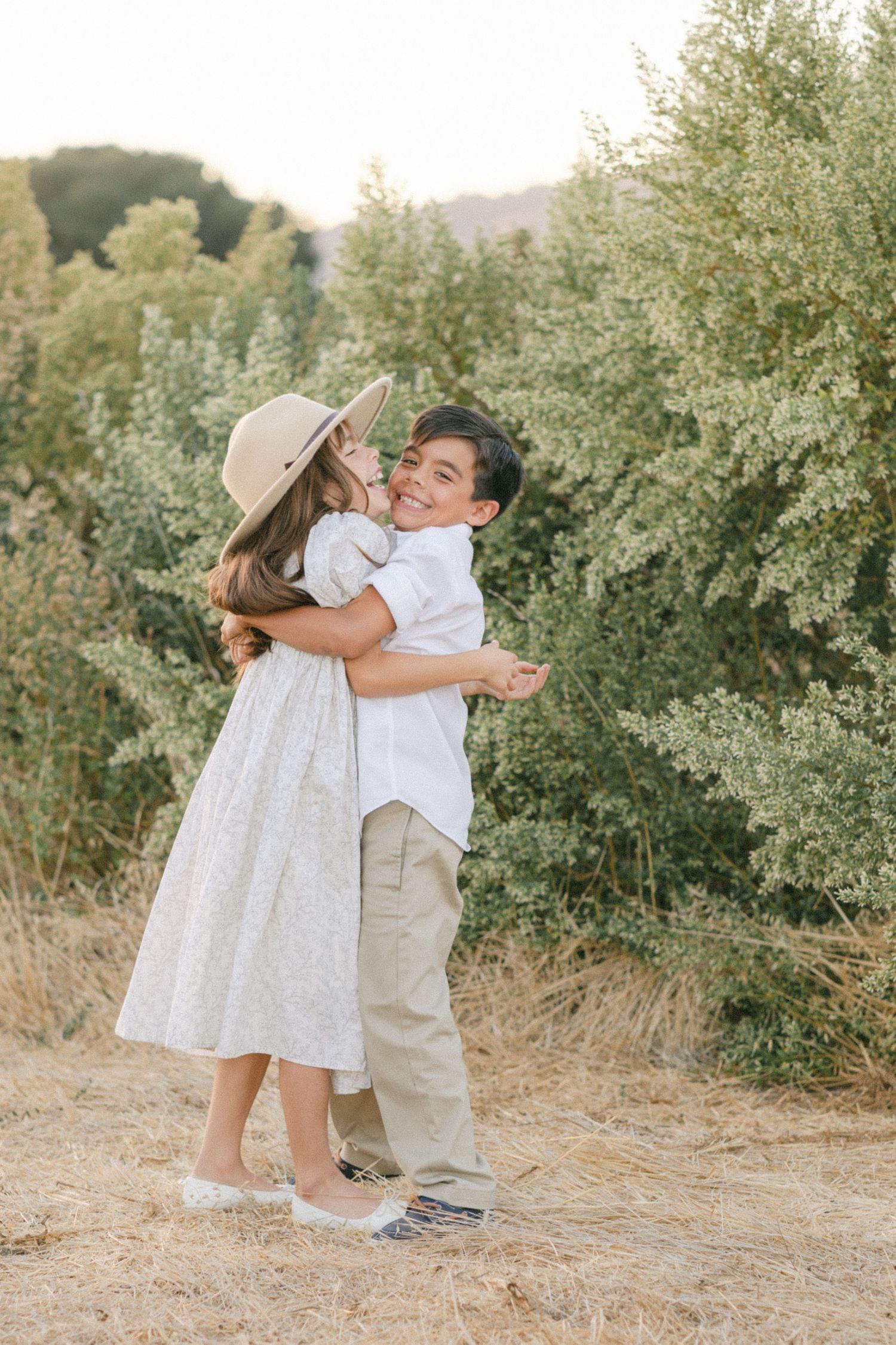Two children hugging outdoors, girl in white dress and sun hat, boy in white shirt and khakis, surrounded by green trees.