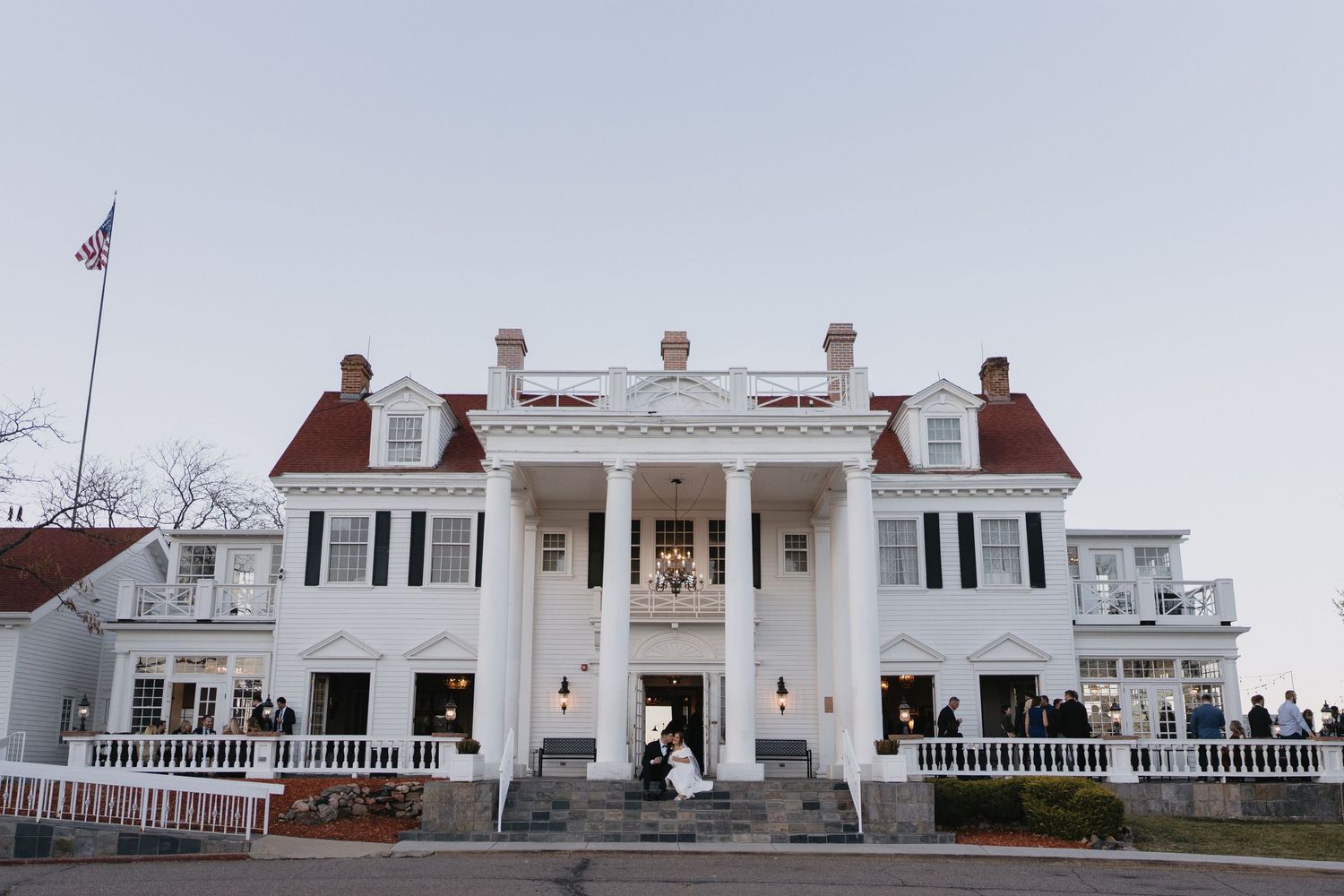 Elegant white colonial mansion wedding venue with grand columns, red roof, and American flag at dusk.