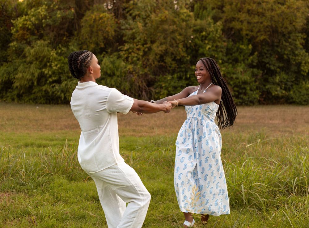 Two people joyfully spinning together in a green meadow, wearing elegant white and floral blue outfits.